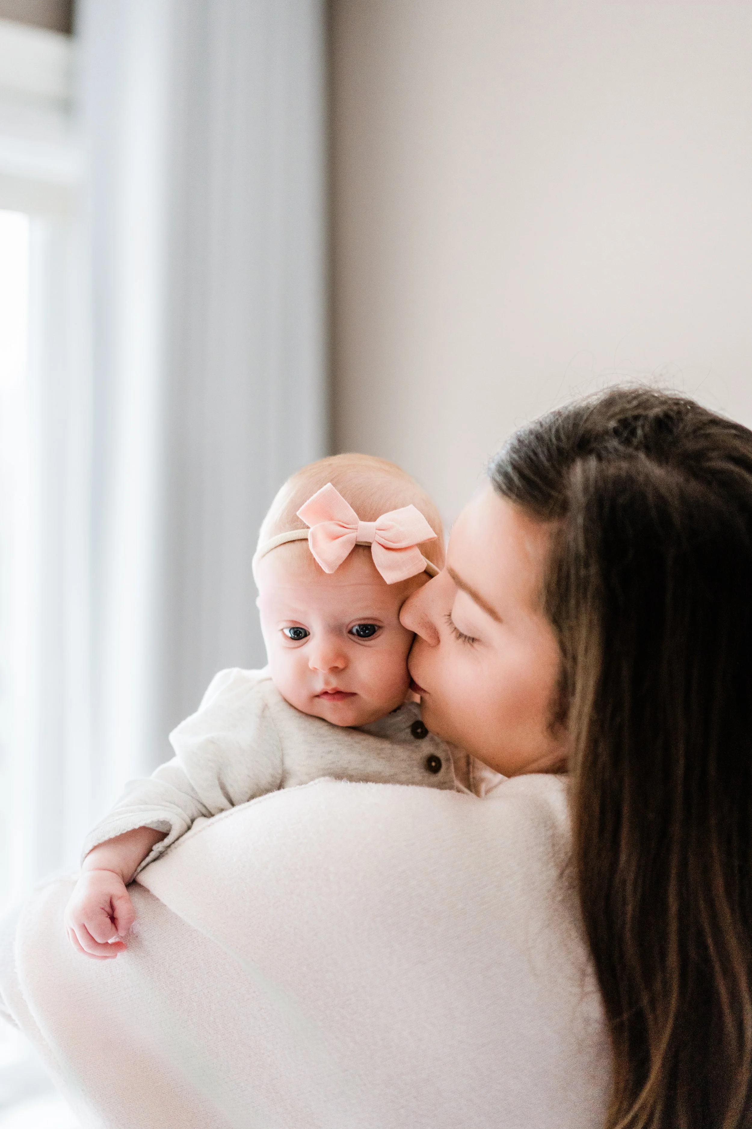 A woman holding a baby girl in her arms. The woman is giving the baby a kiss on the cheek. The baby has a pink bow headband and is looking at the camera.
