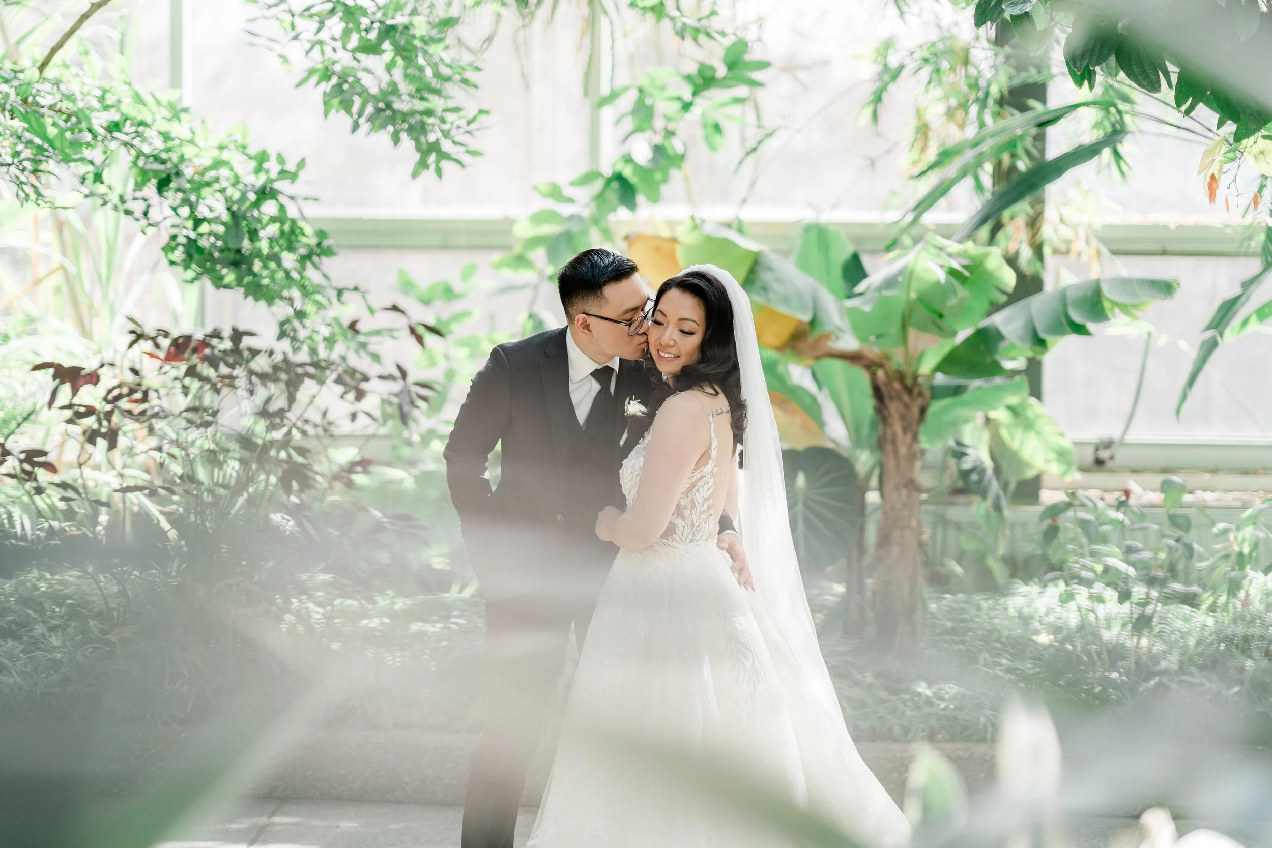 A bride and groom embrace in a lush, green indoor garden, with the groom kissing the bride's cheek. The bride wears a white wedding gown with lace details and veil, while the groom is in a black suit. Sunlight filters through large windows.