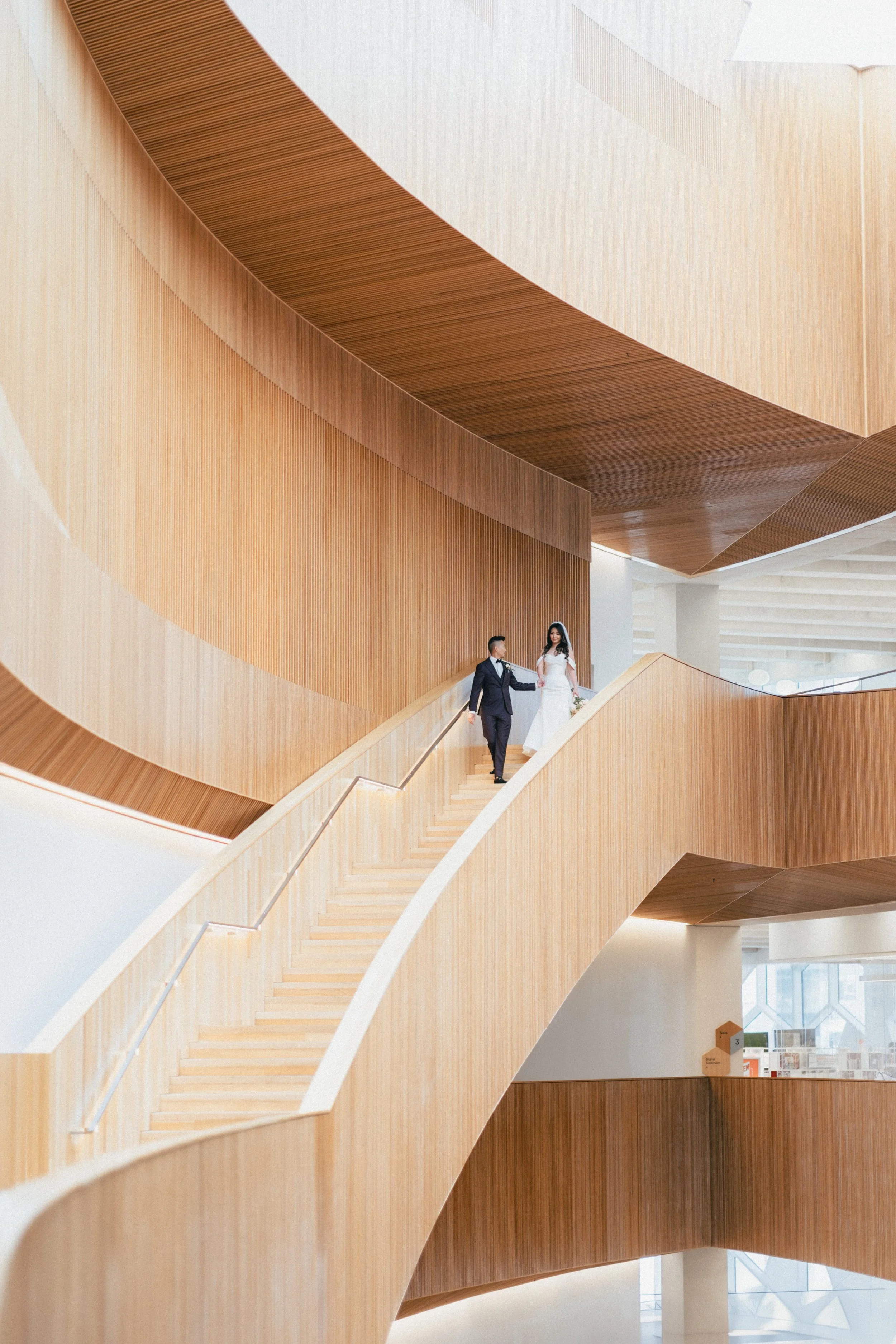 A bride and groom walking down a modern, curved wooden staircase in a spacious, brightly lit building.