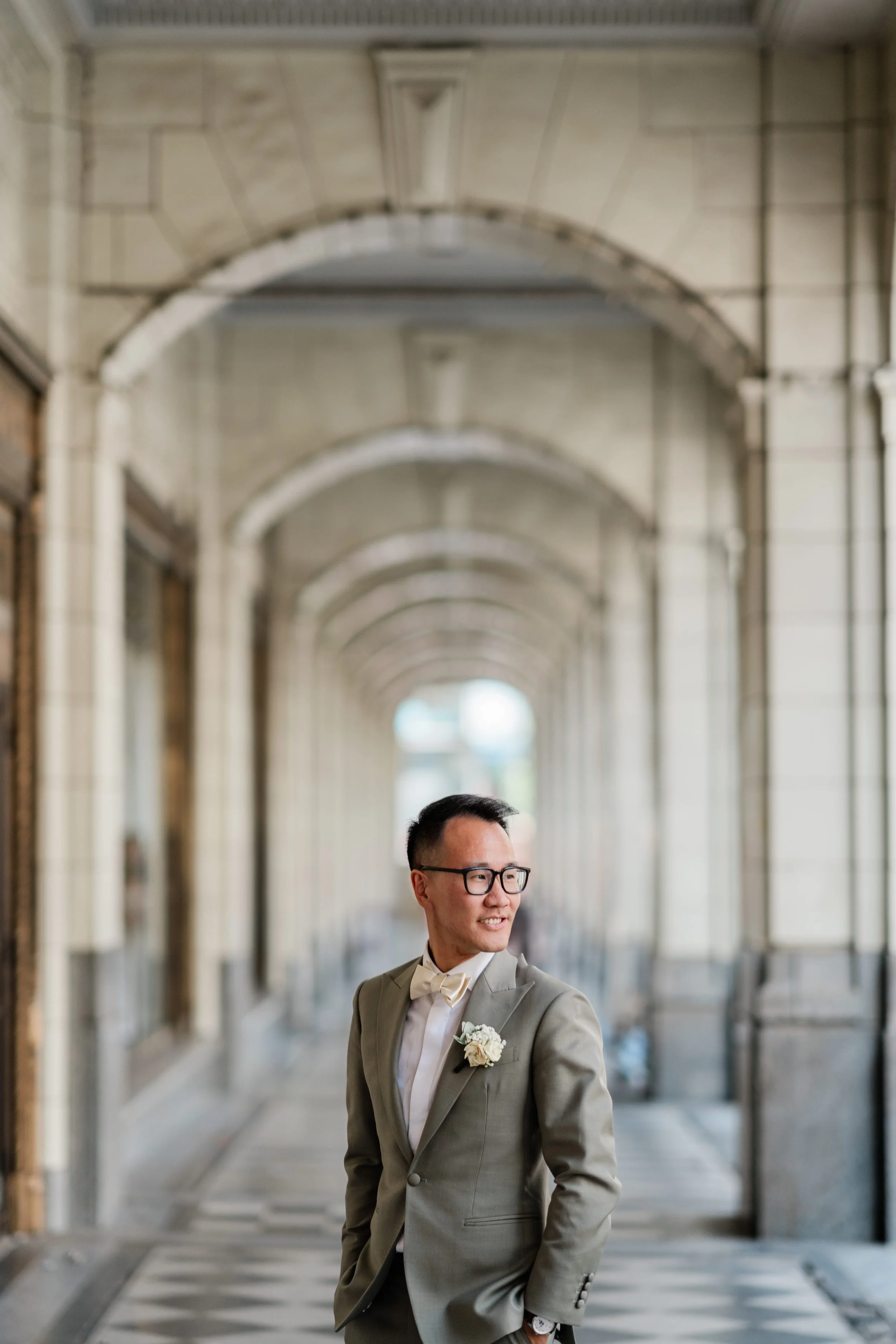A man in a light gray tuxedo and bow tie, wearing glasses and a boutonniere, standing in a historic arched corridor with ornate stonework, looking to his right with a smile.