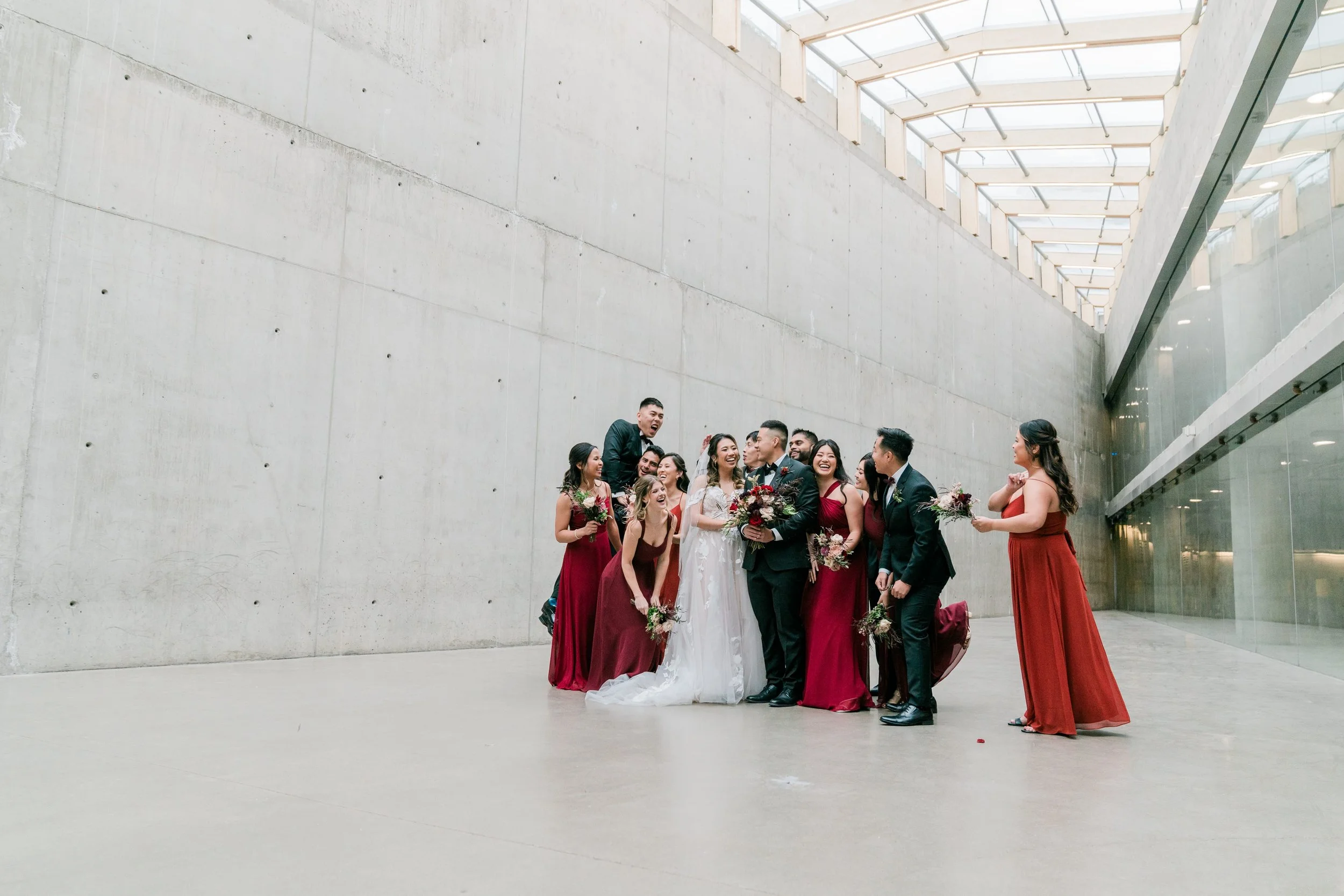 A group of people dressed in formal attire, likely at a wedding, standing in a modern, minimalistic indoor space with concrete walls and large glass windows, celebrating and smiling.