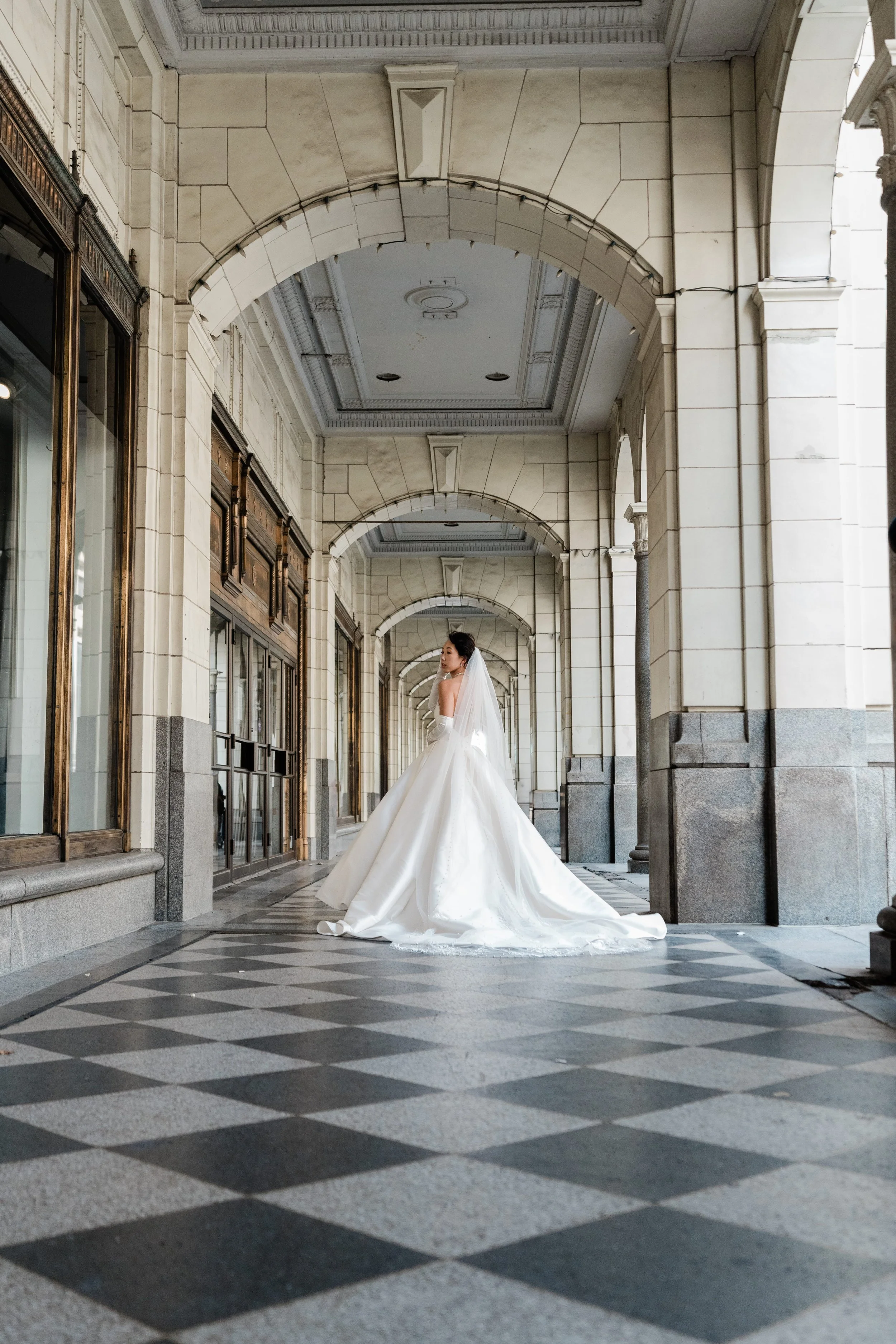 A bride in a white wedding gown and veil standing in an arched corridor with checkered black and white floor and ornate architecture.