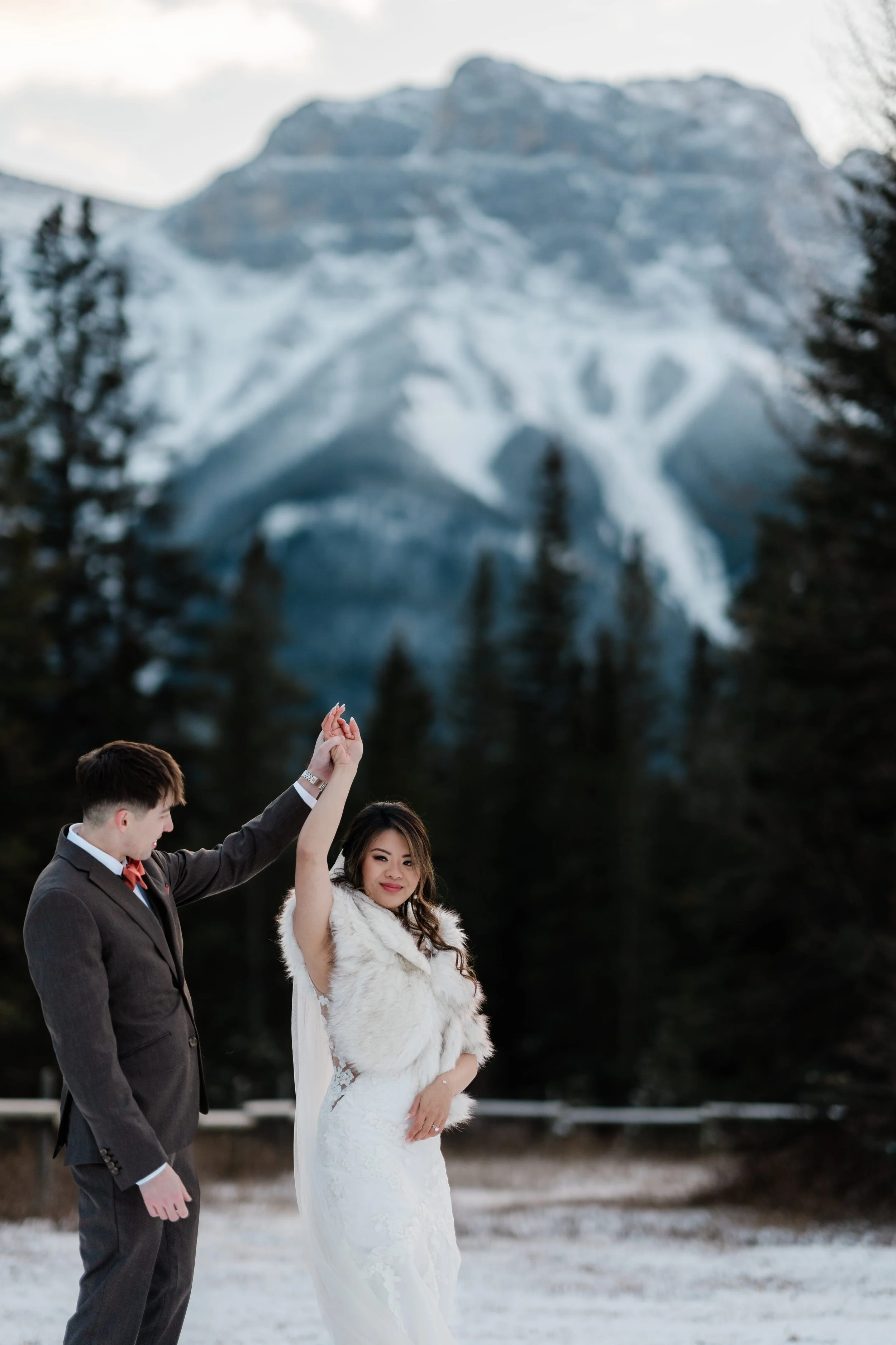 A bride and groom dancing outdoors on snow on a winter day, with a mountain with snow and trees in the background.