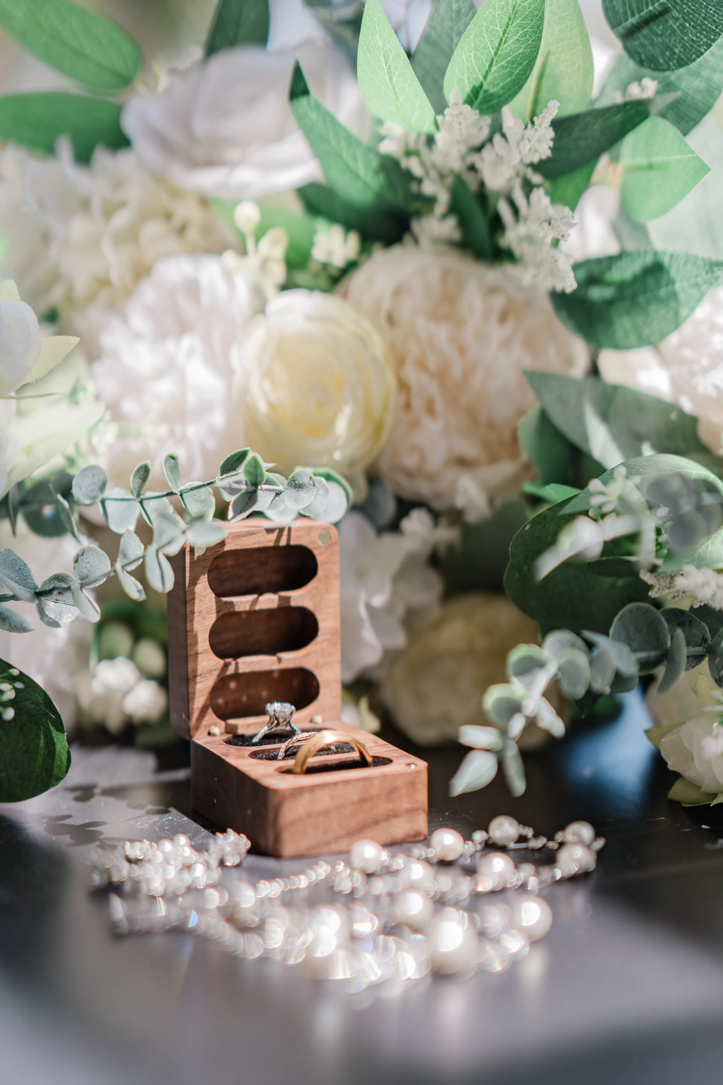 A wooden jewelry box with two rings inside, surrounded by white pearls, and a floral arrangement with green leaves and white flowers in the background.