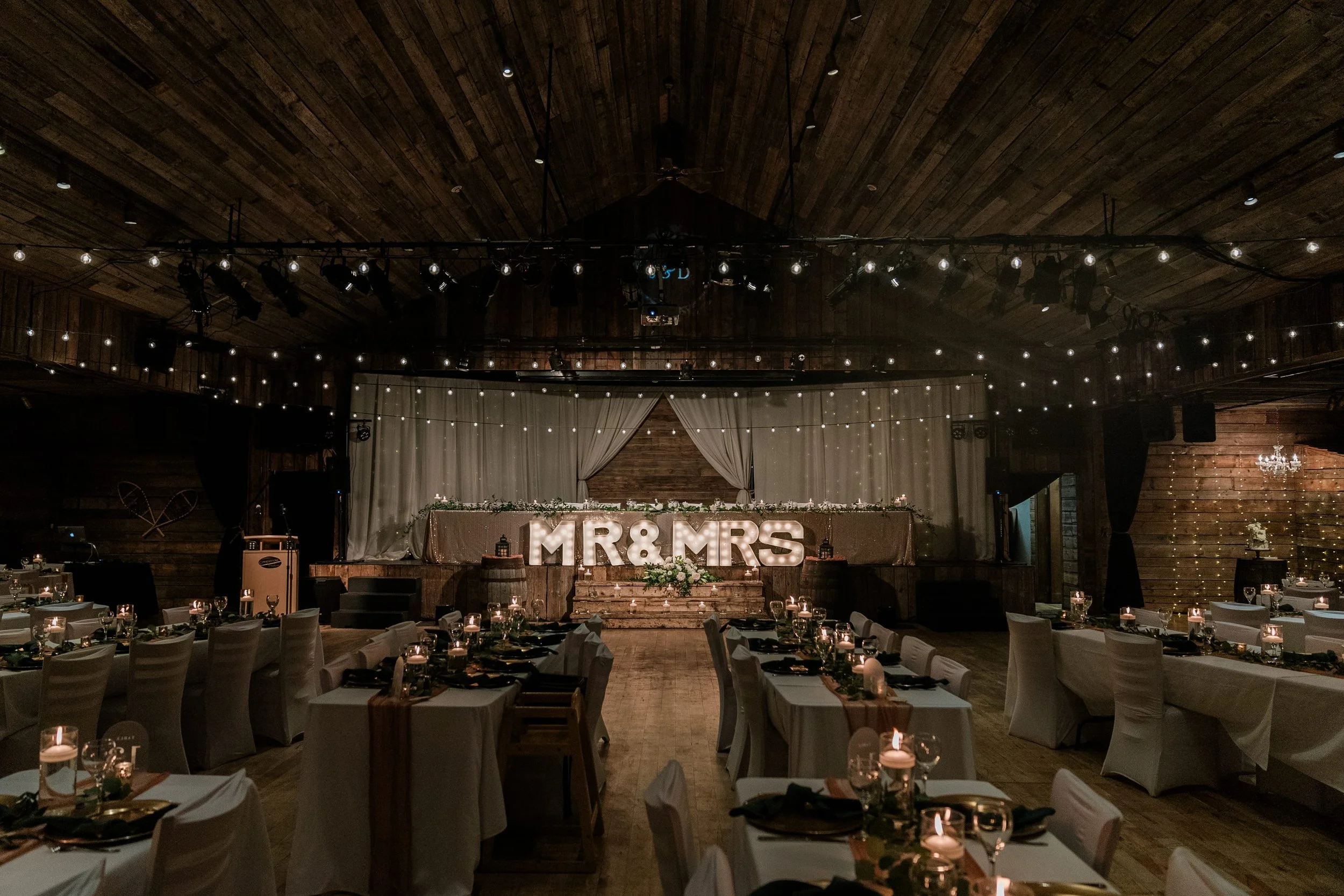 Indoor wedding reception setup with tables decorated with candles and greenery, a stage with large illuminated letters reading "MR & MRS," and wood-paneled ceiling and walls with string lights and drapes.