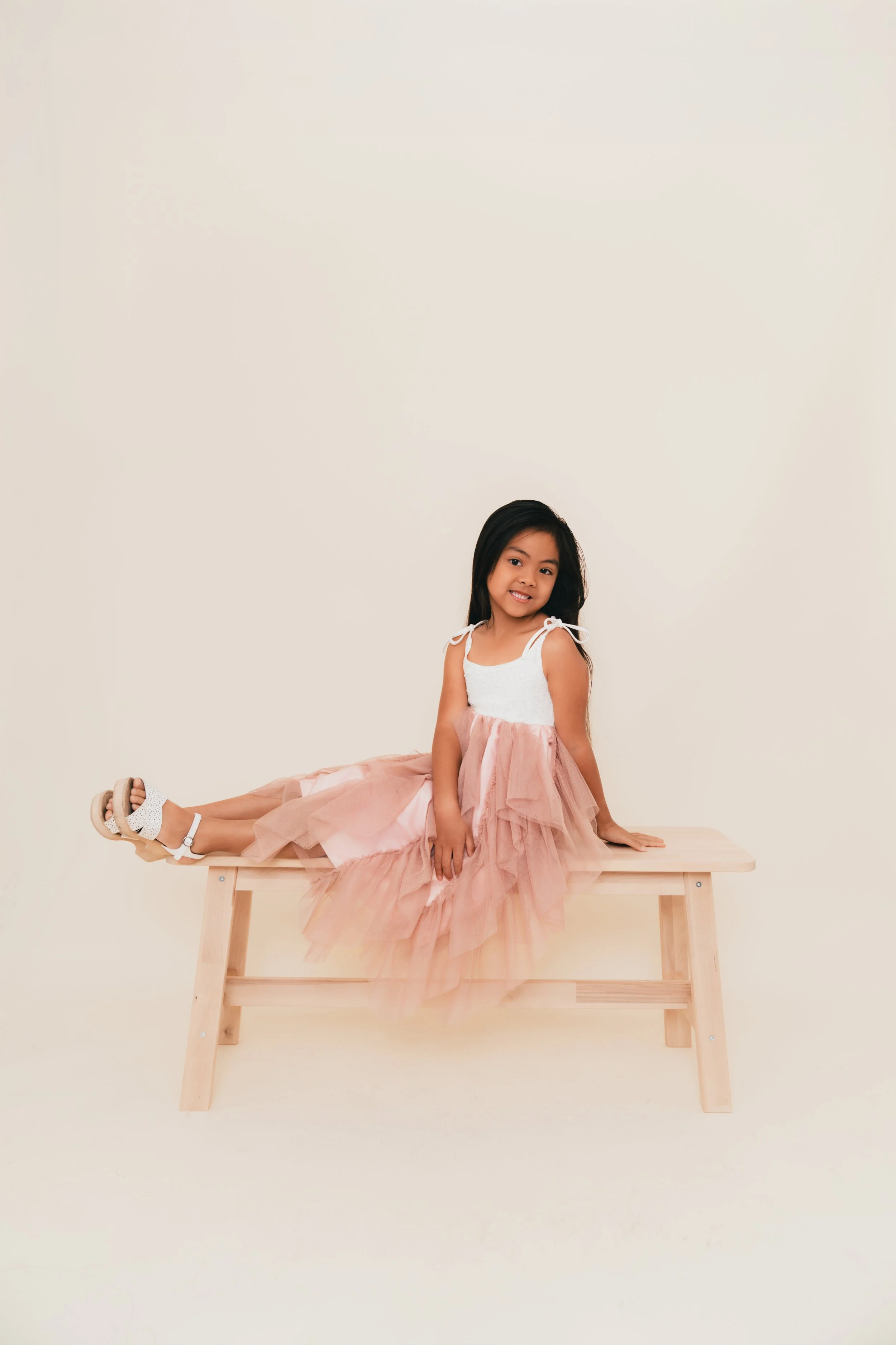 A young girl sitting on a wooden bench in a pink and white dress, smiling at the camera against a plain cream background.