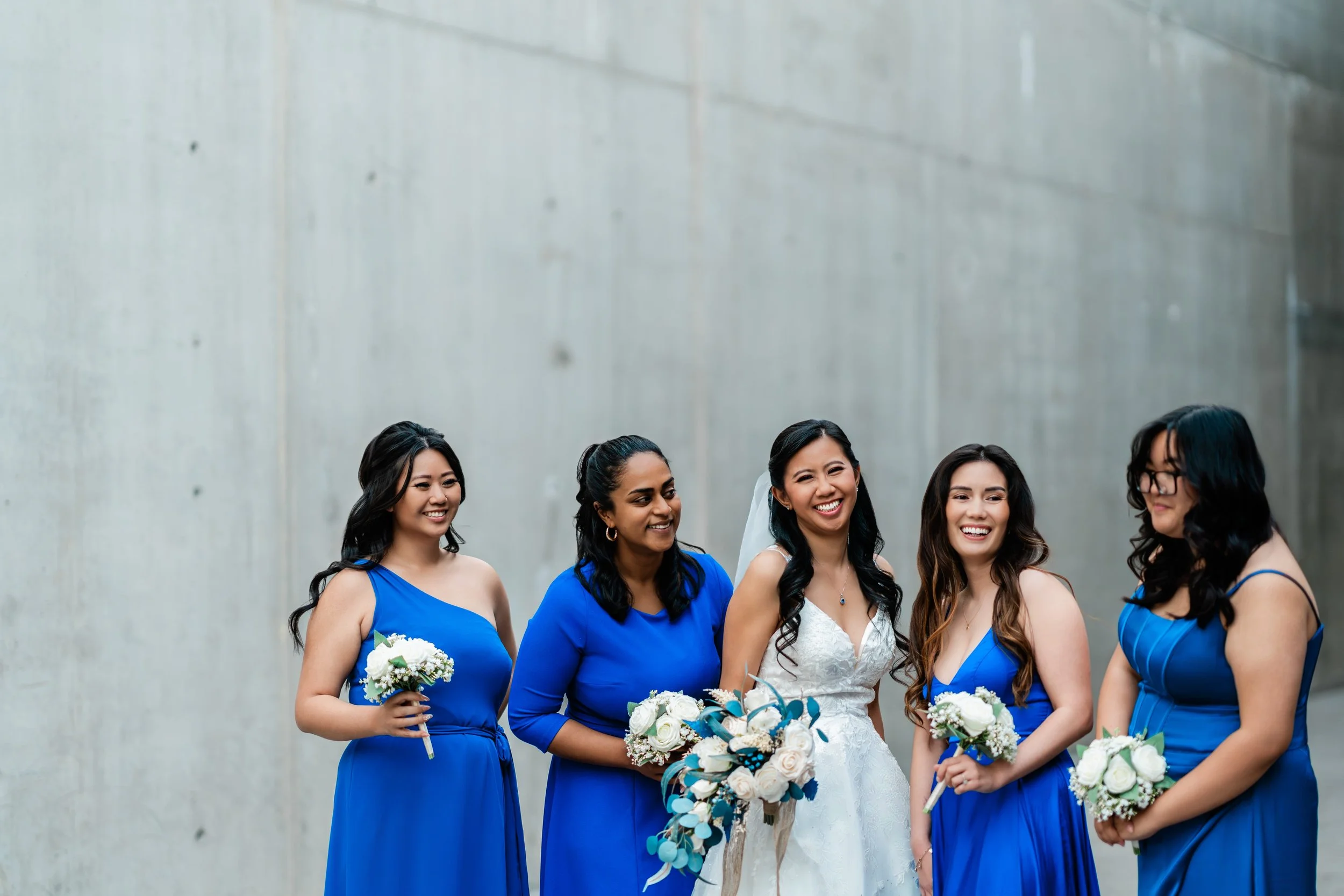 Bride and five bridesmaids smiling, holding bouquets, standing against a plain wall.