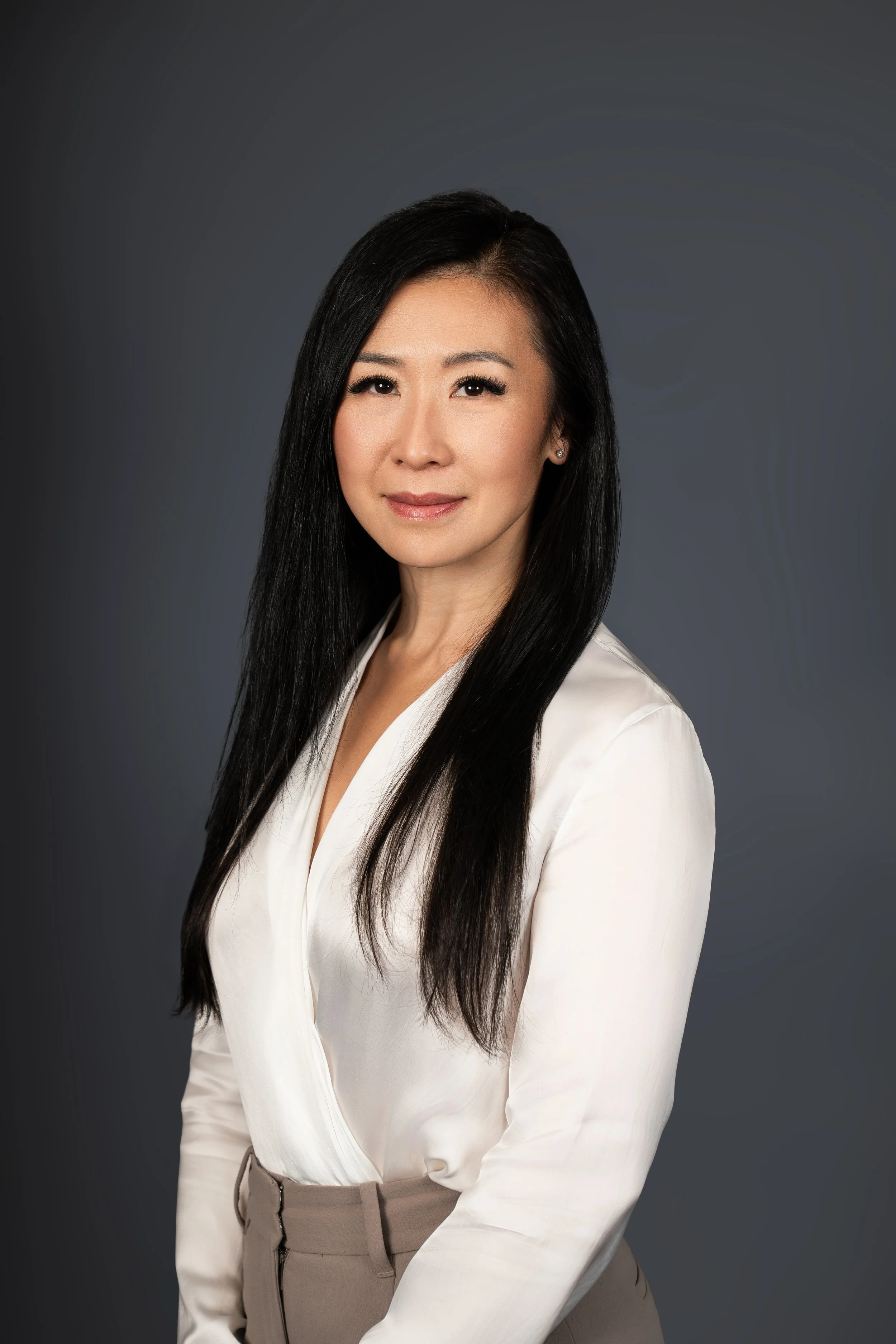 Professional woman with long black hair wearing a white blouse and beige pants, posing against a dark gray background.