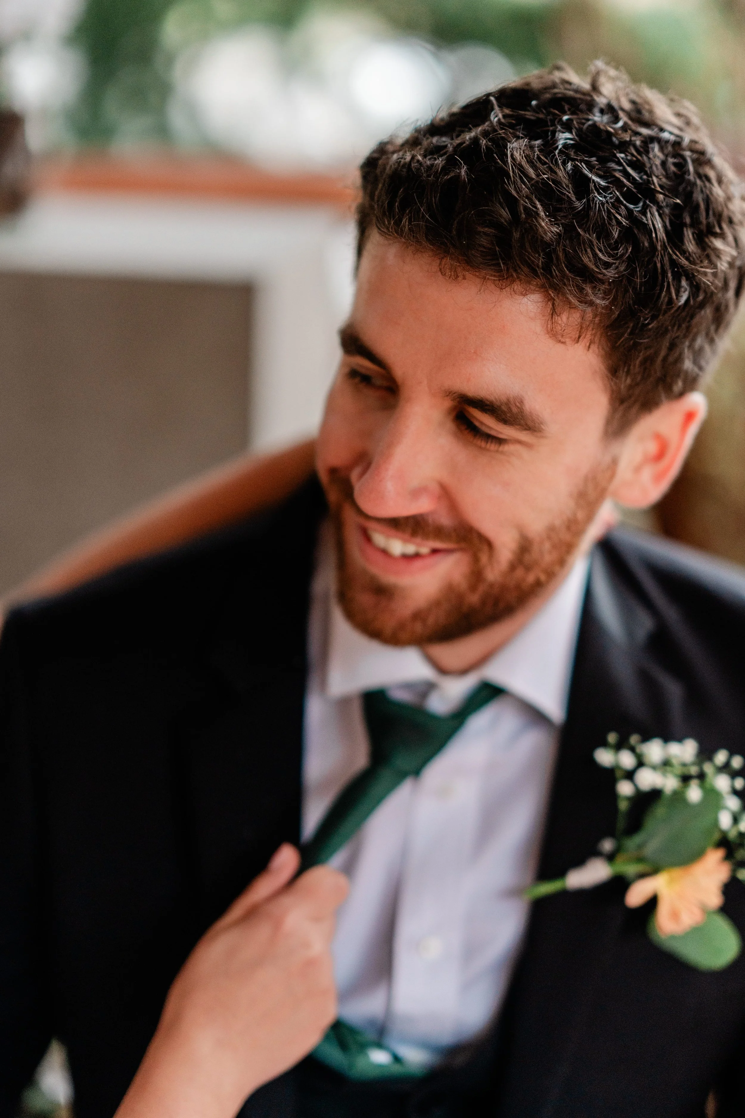 A man in a suit with a boutonnière, smiling as someone adjusts his green tie.