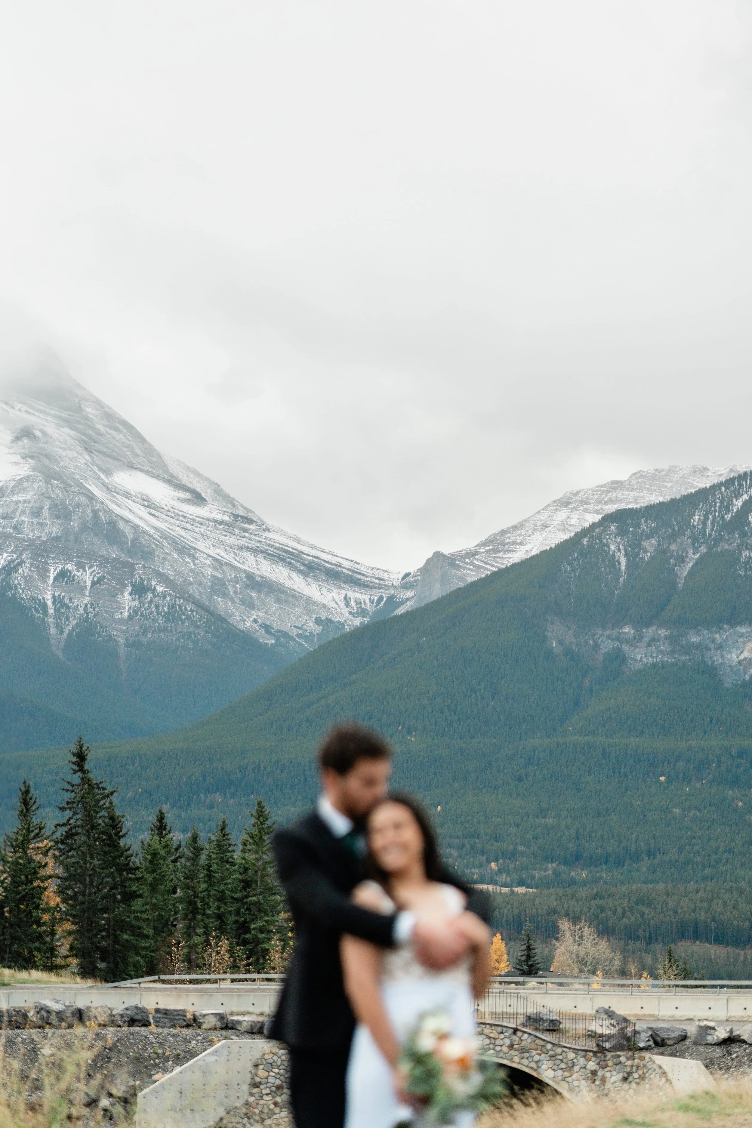 A blurred couple in wedding attire standing outdoors with mountains and trees in the background.