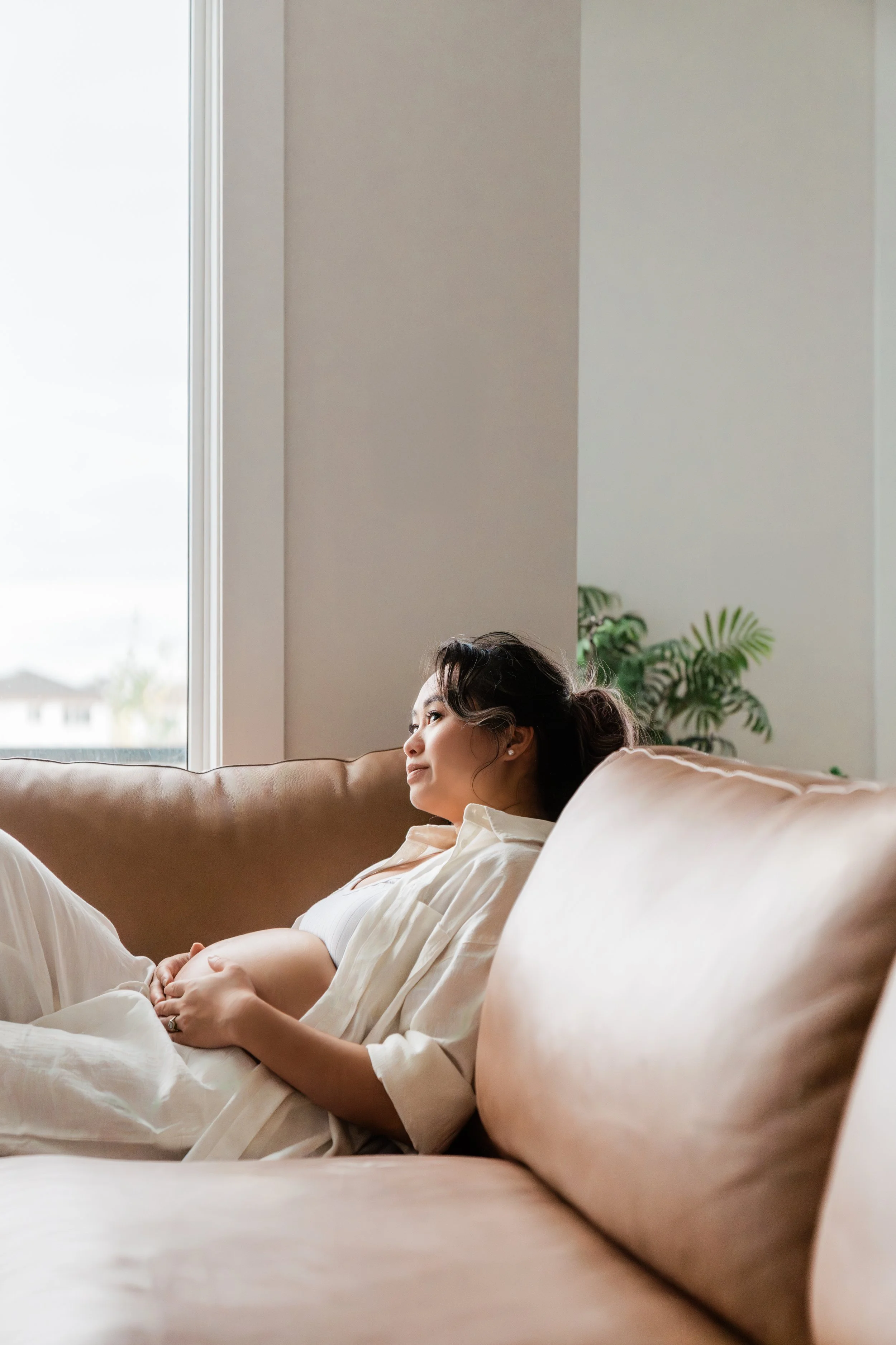 Pregnant woman relaxing on a beige sofa next to a window, with houseplants in the background.