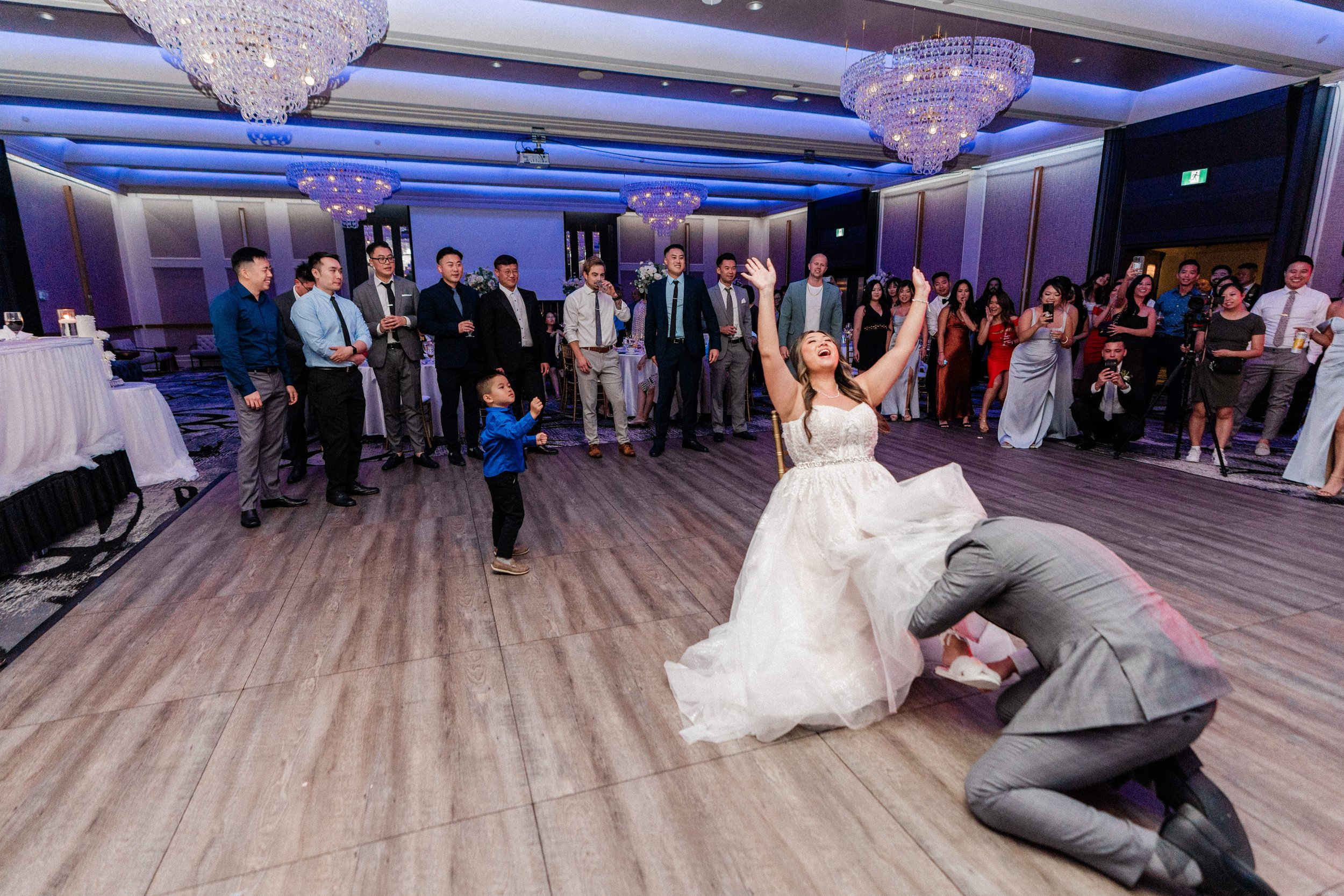 A bride in a white wedding dress is dancing joyfully with her arms raised in a wedding reception hall, surrounded by guests watching and taking photos, with a man kneeling in front of her and a small boy nearby.