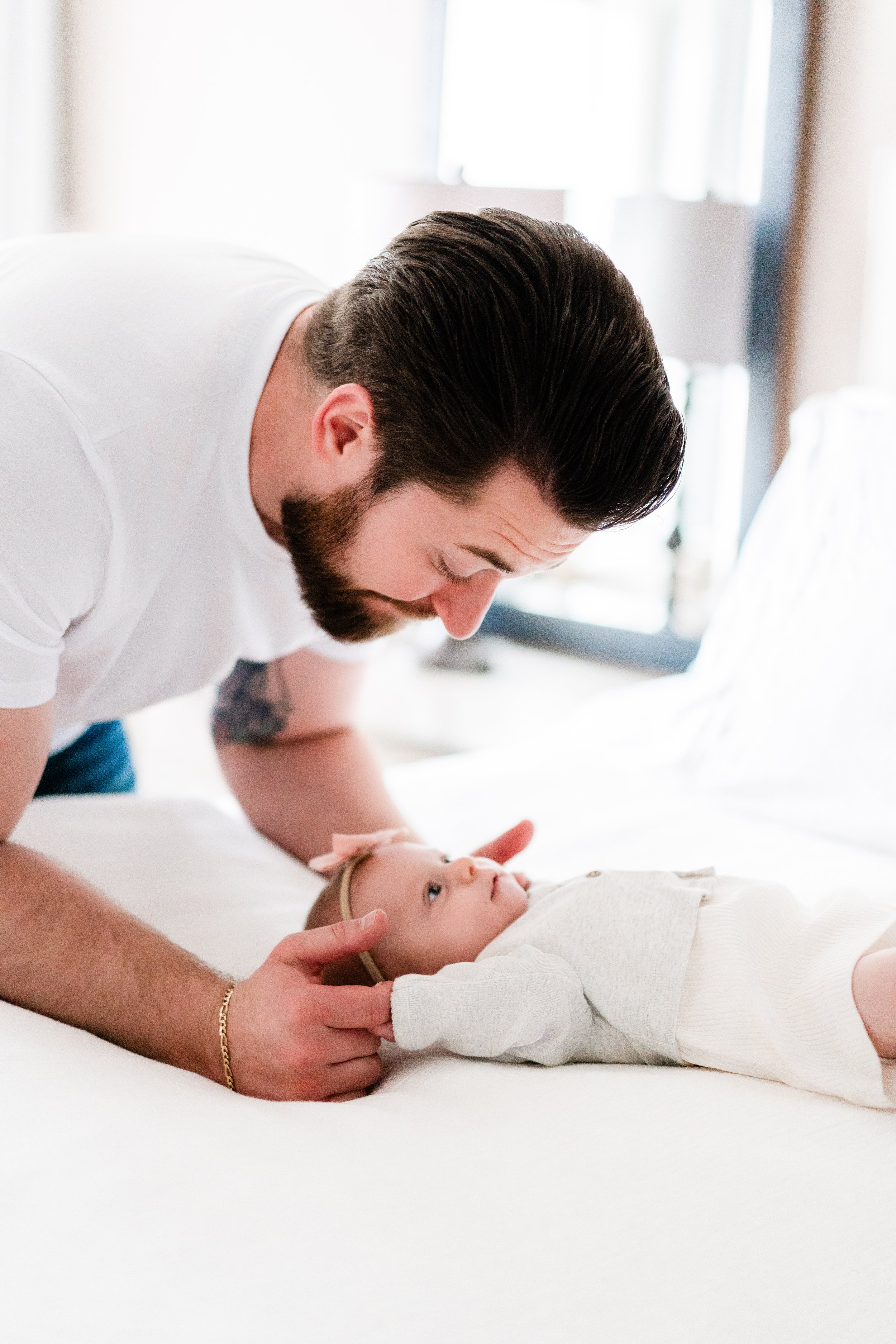 A man with dark hair and a beard is leaning over a baby girl who is lying on a bed, holding her hand and looking at her affectionately in a bright room.