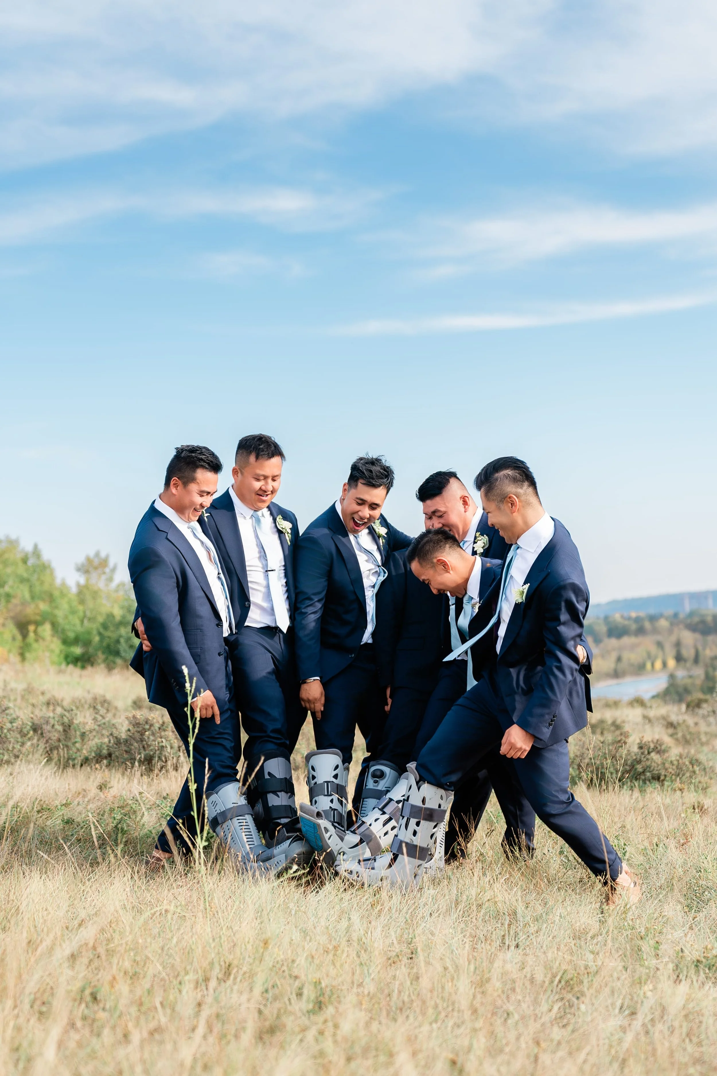 Seven men dressed in suits standing outdoors in a field, looking down at a group of medical exoskeletons on the ground, all smiling and looking amused.