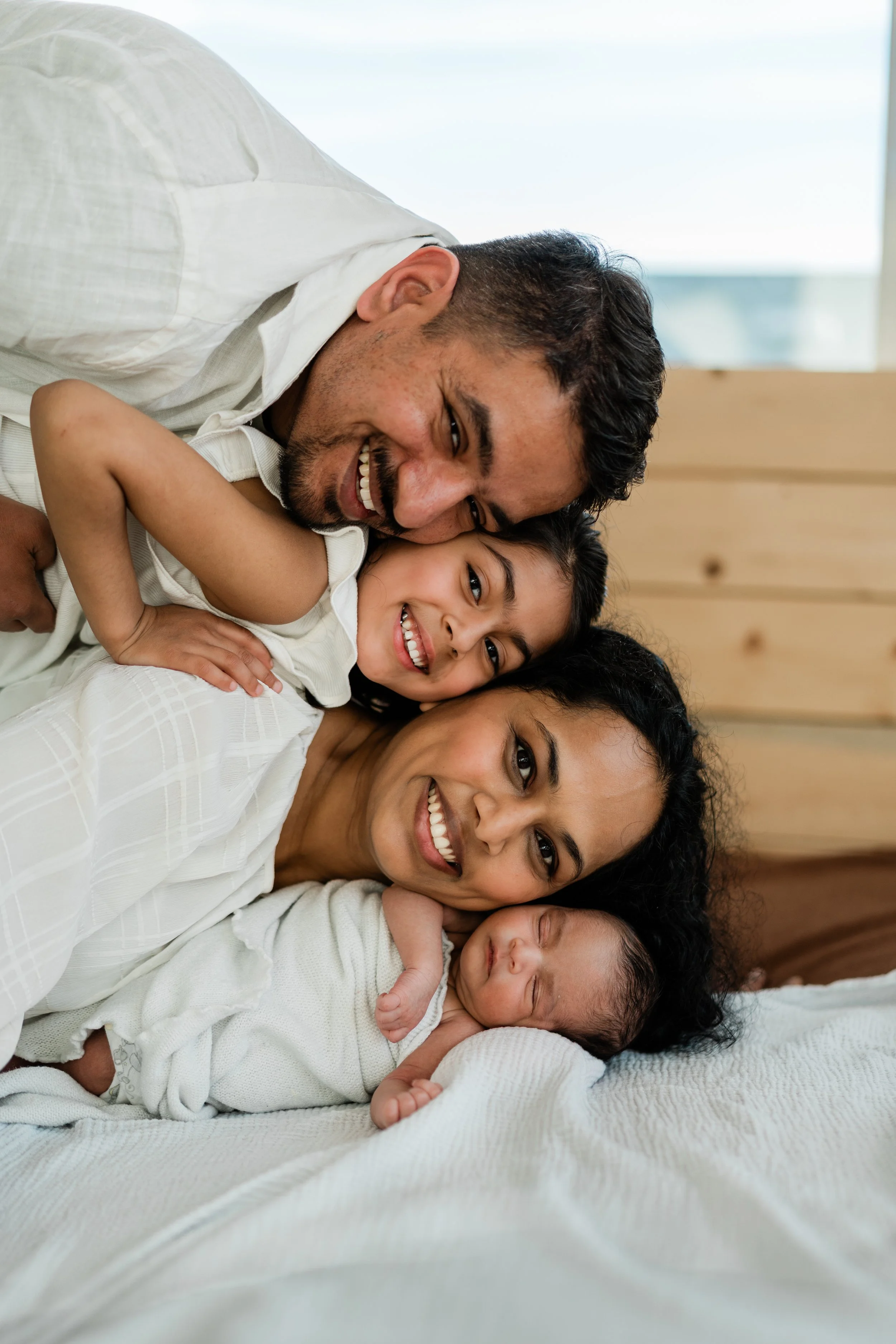 Family of five lying on bed, smiling and hugging, with a newborn baby, in a bright room with wooden wall and large window.