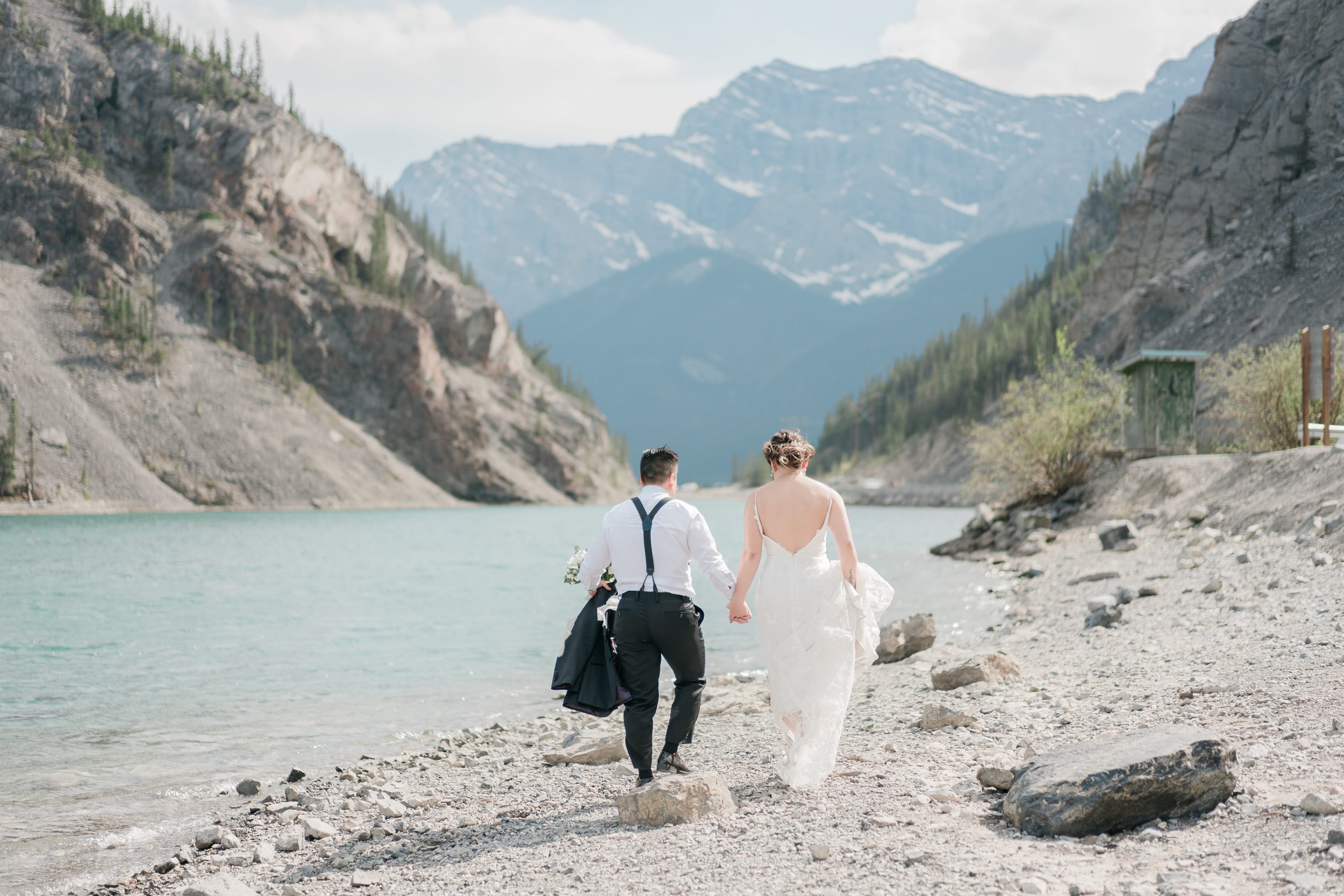 A couple walking hand in hand along a rocky shoreline by a turquoise lake surrounded by mountains and trees, with snowy peaks in the distance. The woman is in a white wedding dress, and the man is in a white shirt with suspenders and black pants, car