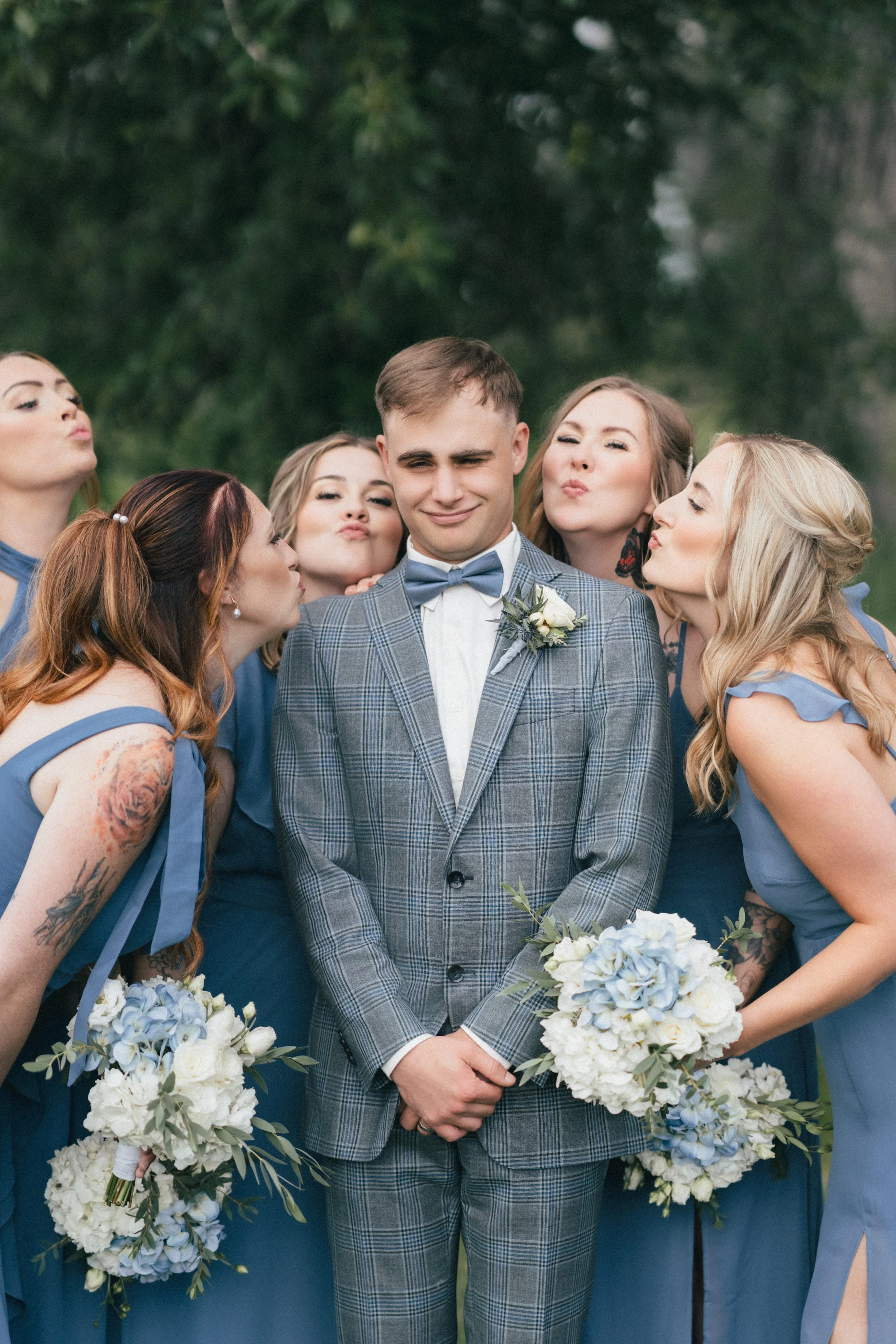 A groom in a gray plaid suit and blue bow tie standing with five women in blue bridesmaid dresses, all holding bouquets, outside with green trees in the background. The women are kissing or puckering their lips toward the groom.