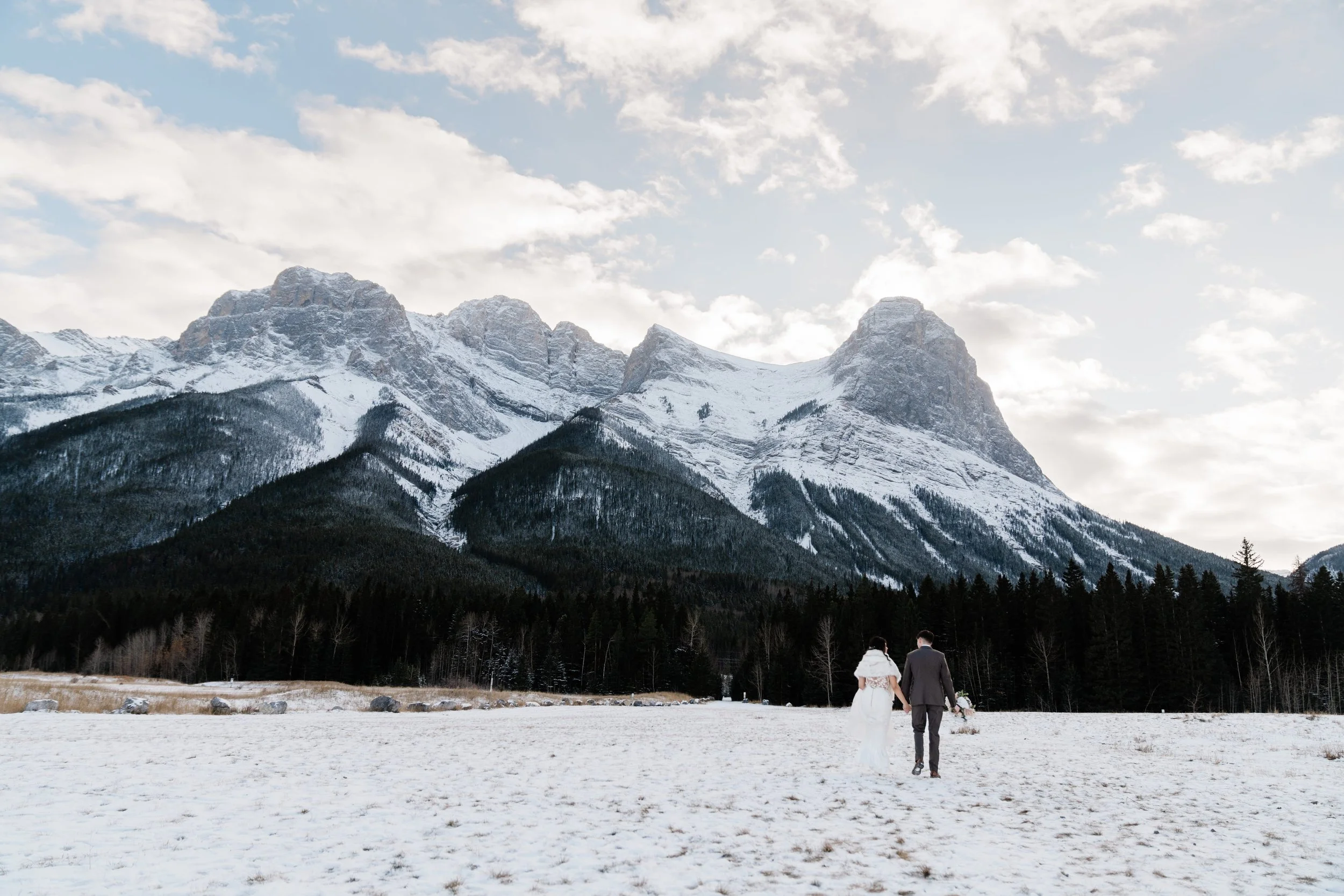 A couple walking hand in hand in a snow-covered field with a mountain range in the background and a partly cloudy sky overhead.