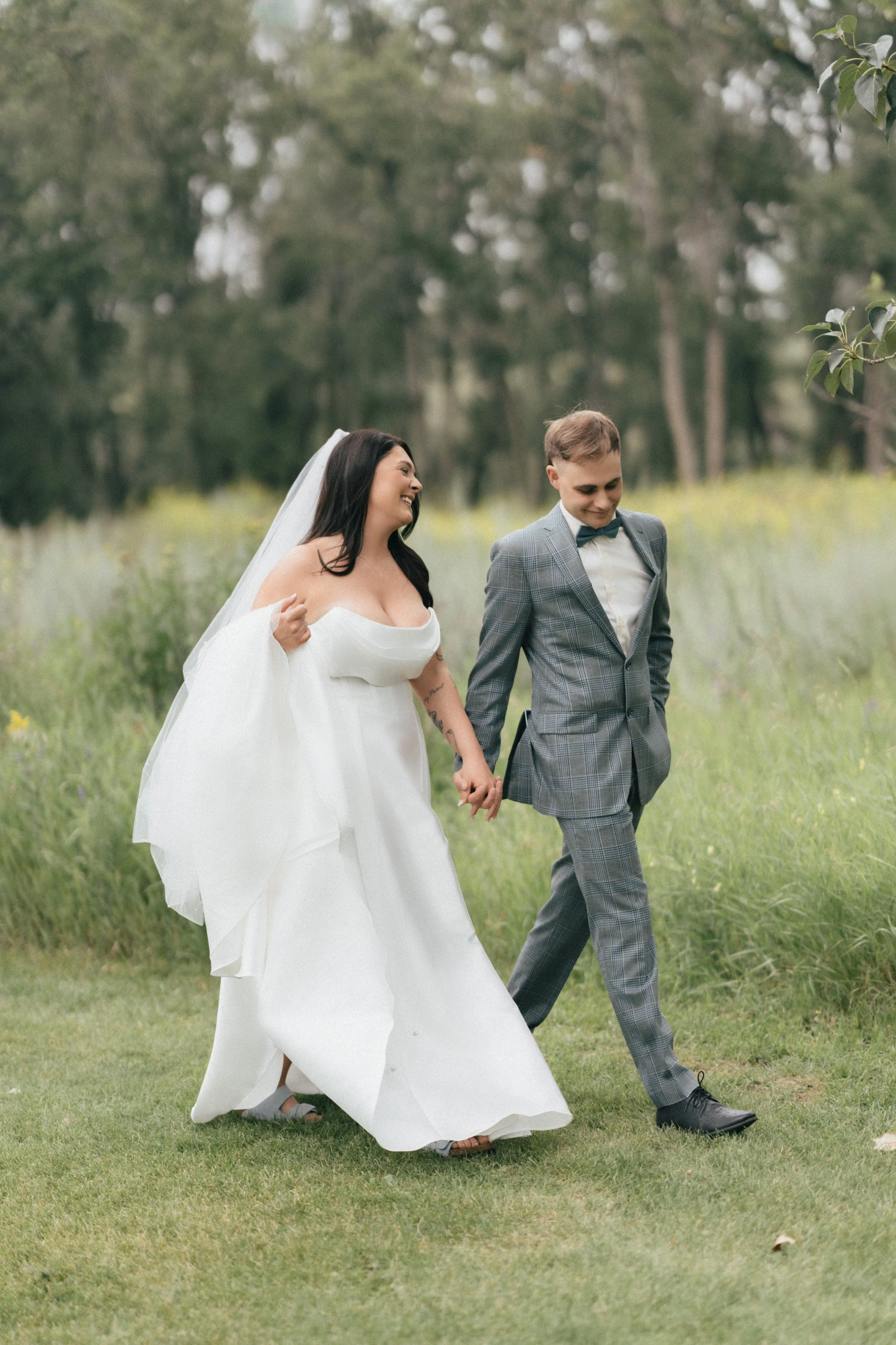 A newlywed couple holding hands and walking through a grassy field surrounded by trees, smiling and enjoying their moment together.