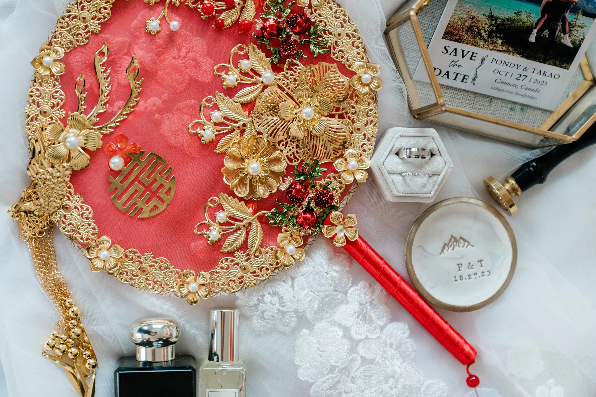 A collection of wedding accessories and decorations, including gold jewelry, a bouquet of red flowers, a ring in a white ring box, perfume bottles, a round decorative item with initials and date, and a framed 