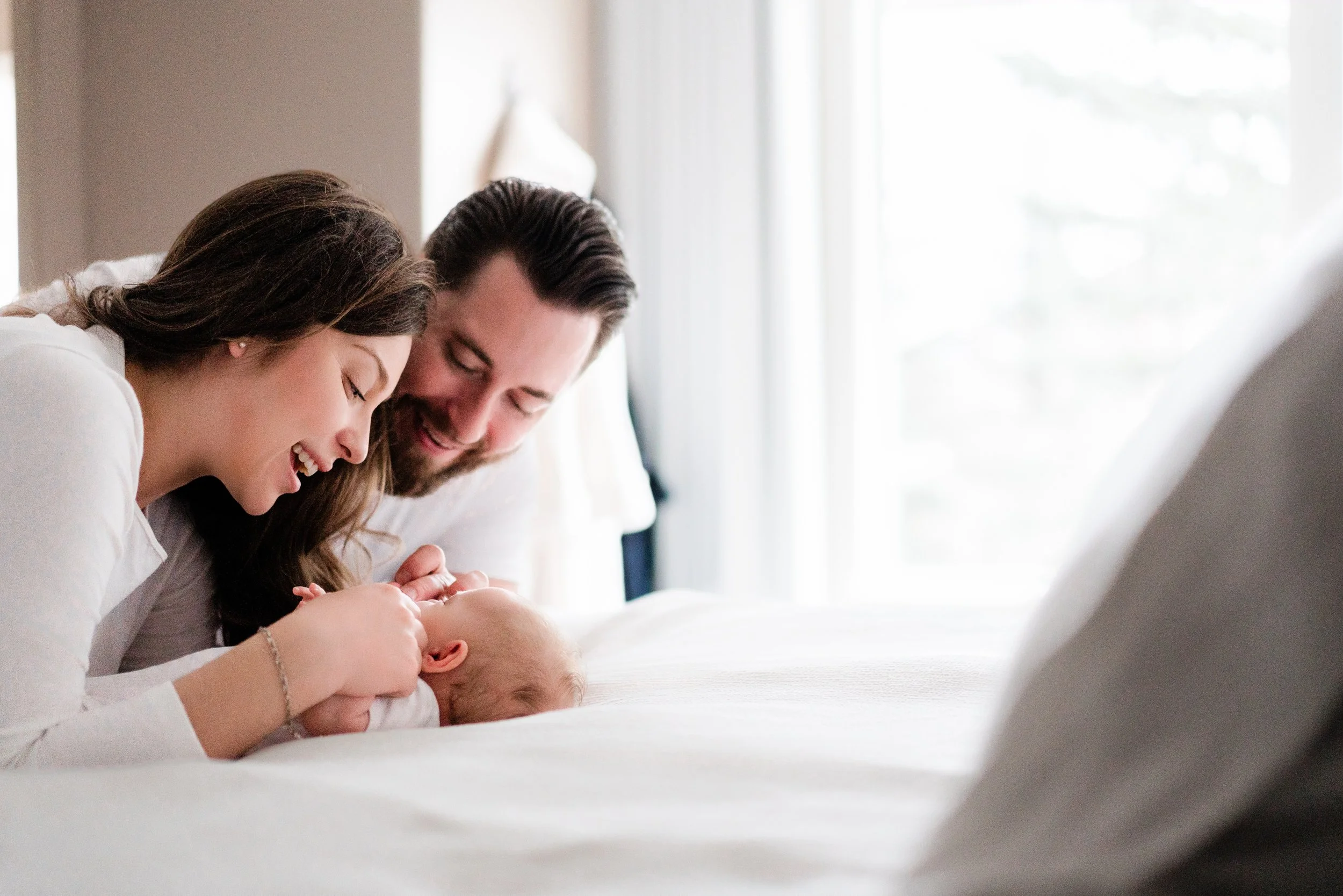 A woman and a man playfully interact with a baby on a bed near a large window, smiling and enjoying a tender moment together.
