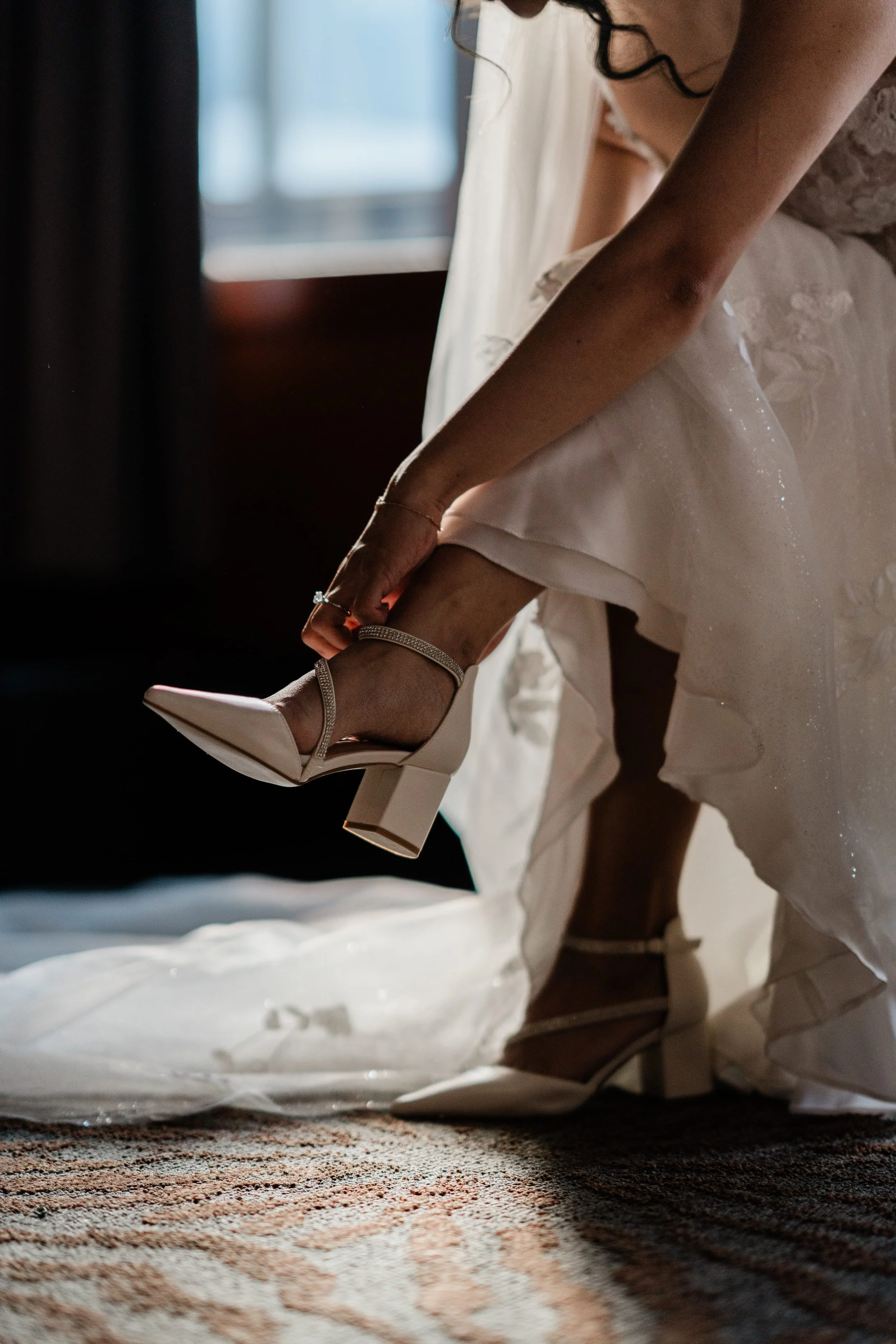 A bride is putting on a white high-heeled shoe with ankle straps, while kneeling on a patterned carpet. She is wearing a wedding dress, and the light coming from a window illuminates her and the surroundings.