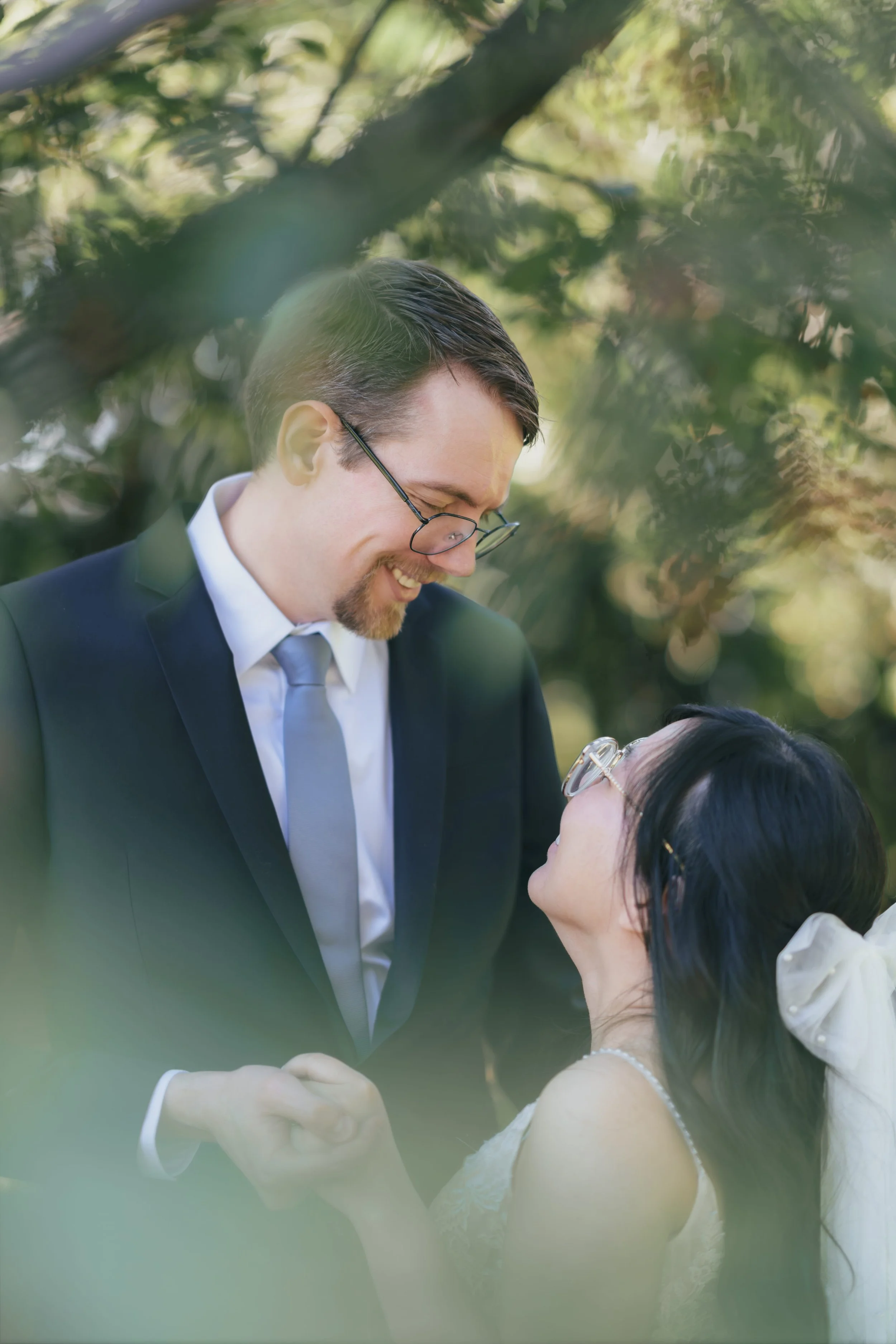 A newlywed couple sharing a moment outdoors: a man in a suit and glasses smiling at a woman in a wedding dress, surrounded by trees.