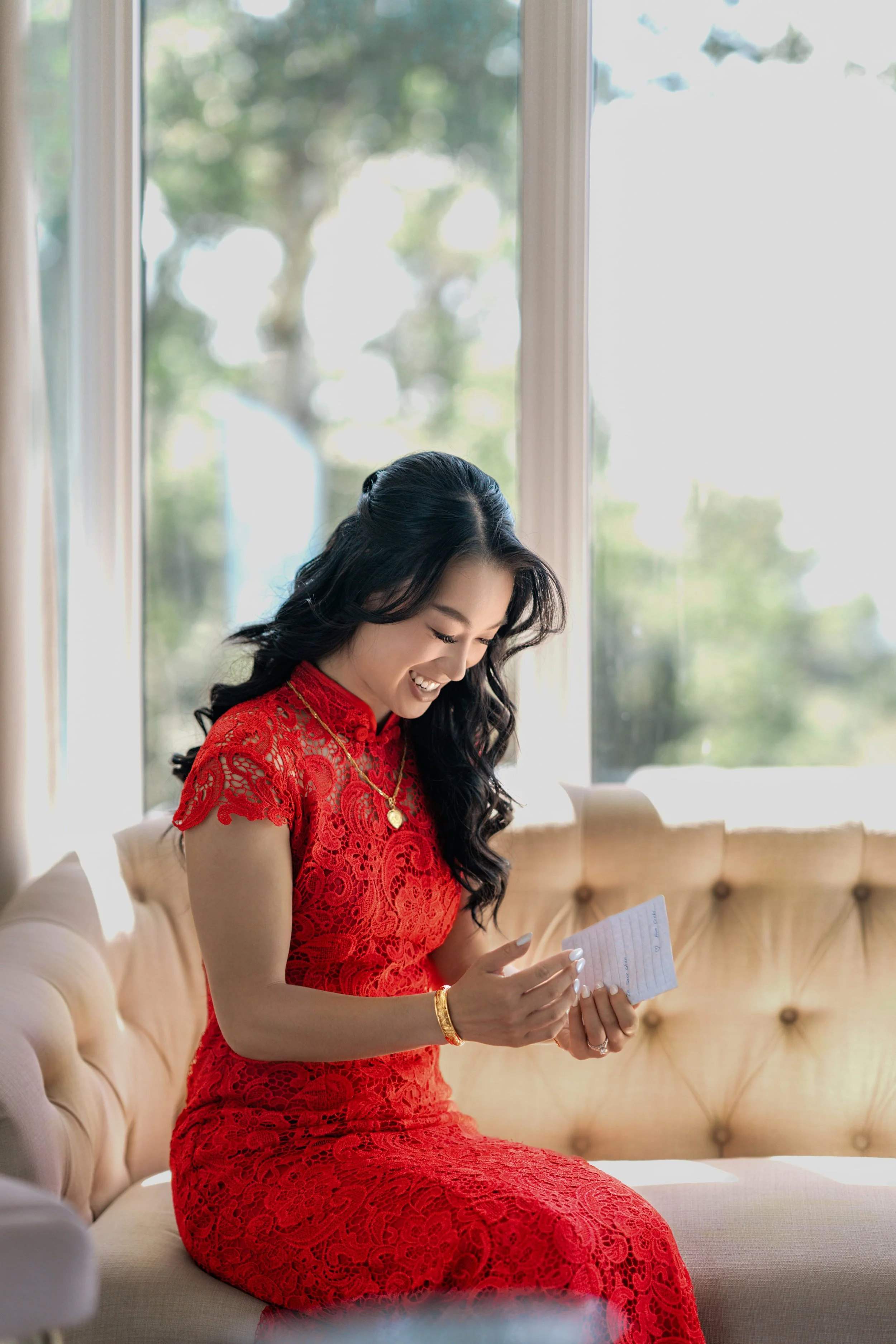 A woman in a red lace dress sitting on a beige couch, smiling and looking at a small piece of paper, with large windows and green trees in the background.