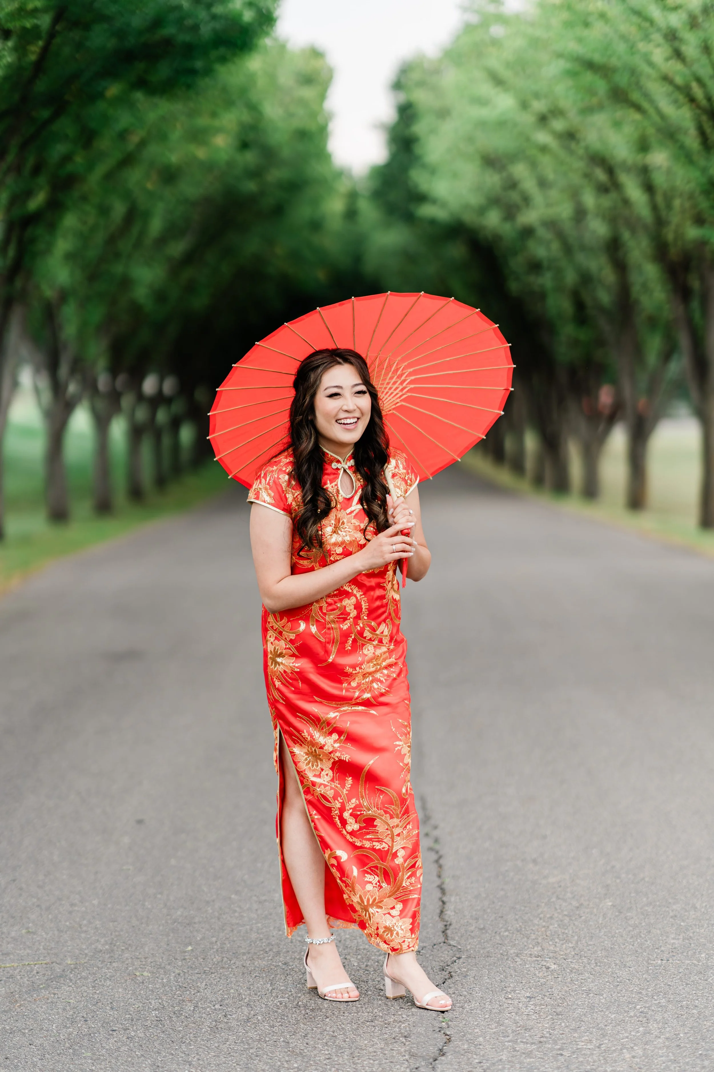 A woman smiling and holding a red umbrella while walking on a tree-lined road, wearing a red and gold traditional dress.