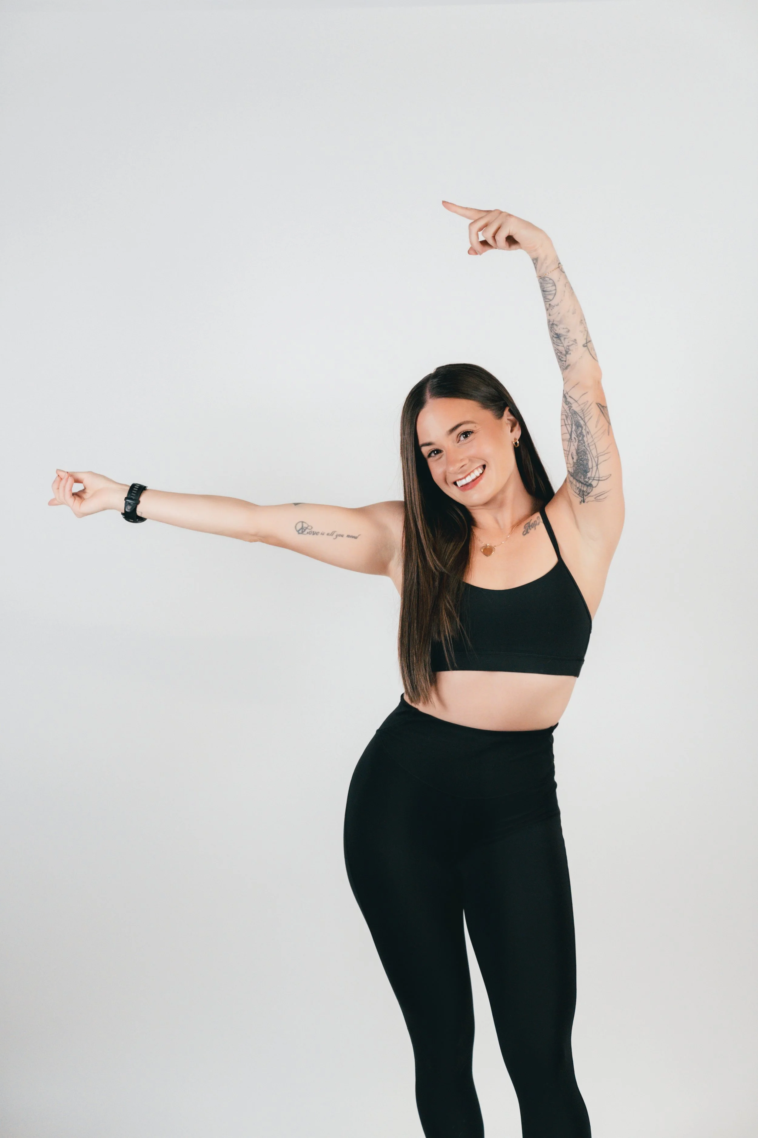 Smiling woman in black workout outfit stretching with arms raised against plain white background.