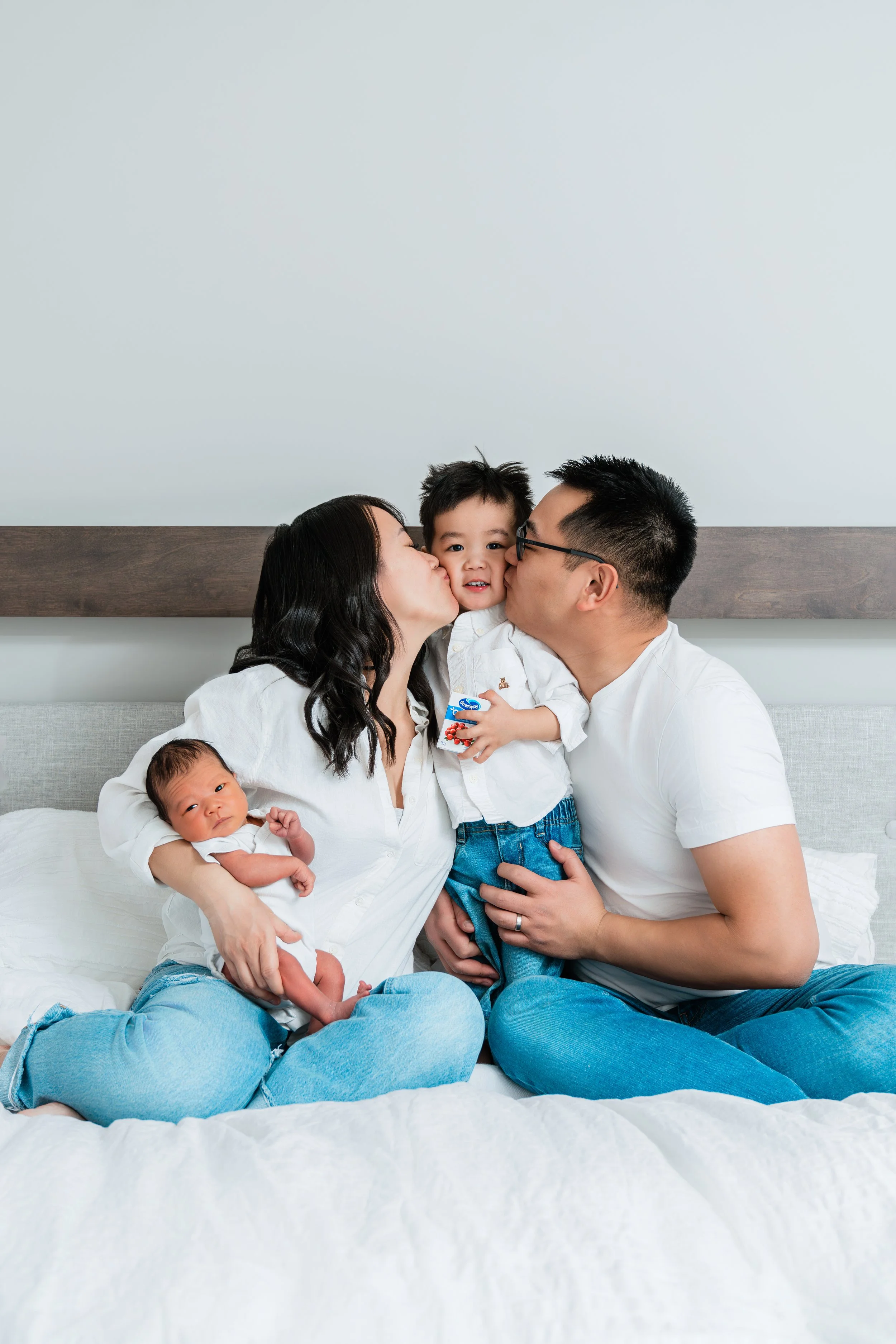 A family of four with two young children, a boy and a baby, sitting on a bed and sharing a moment of affection. The mother and father are kissing the boy on the cheeks, and the baby is lying on the mother’s lap looking at the camera.