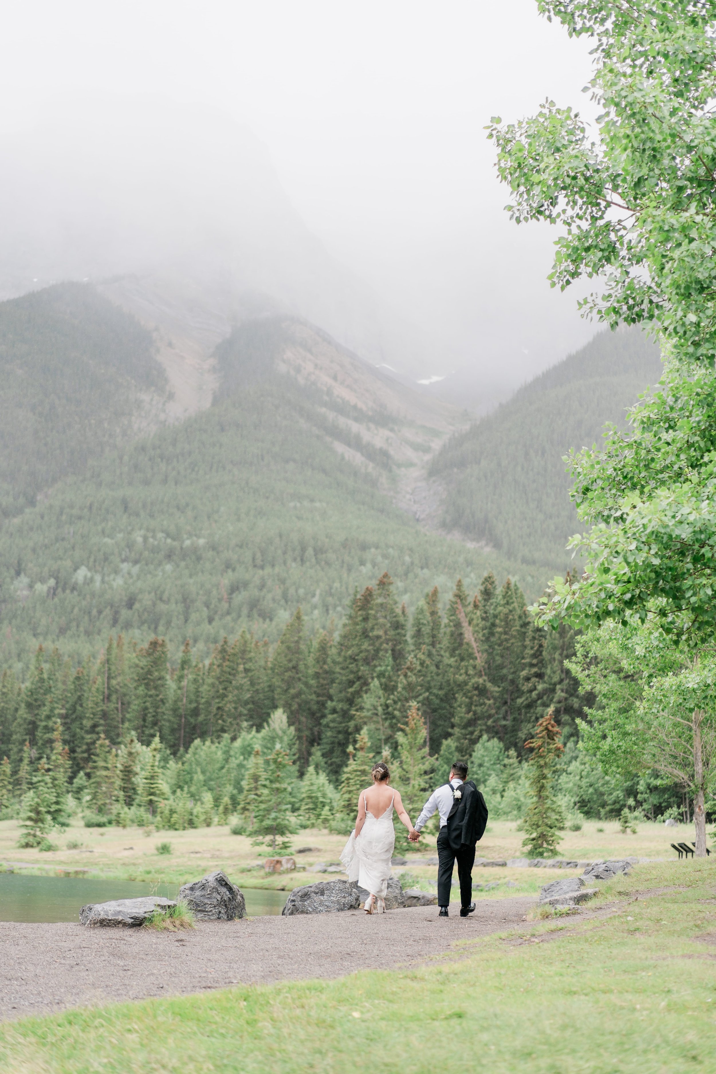 A bride and groom walking hand-in-hand along a lakeshore with a backdrop of forested mountains and a cloudy sky.