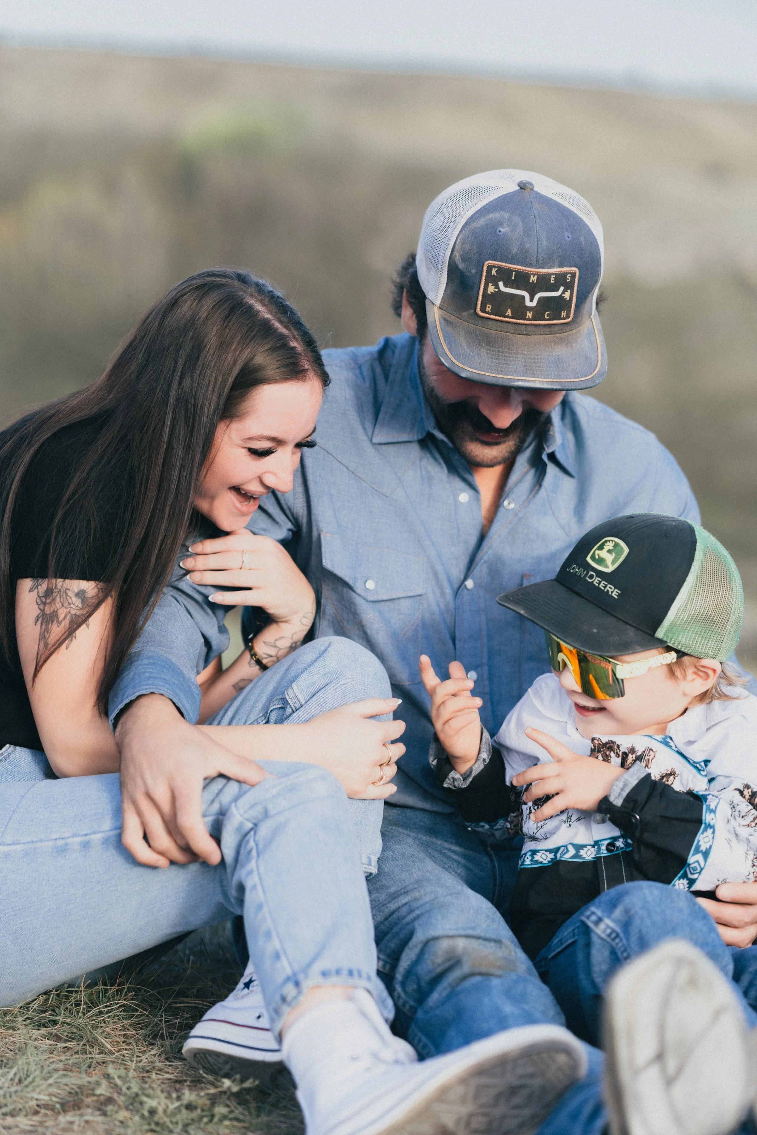 A family of three sitting outdoors on the grass, smiling and interacting with each other.