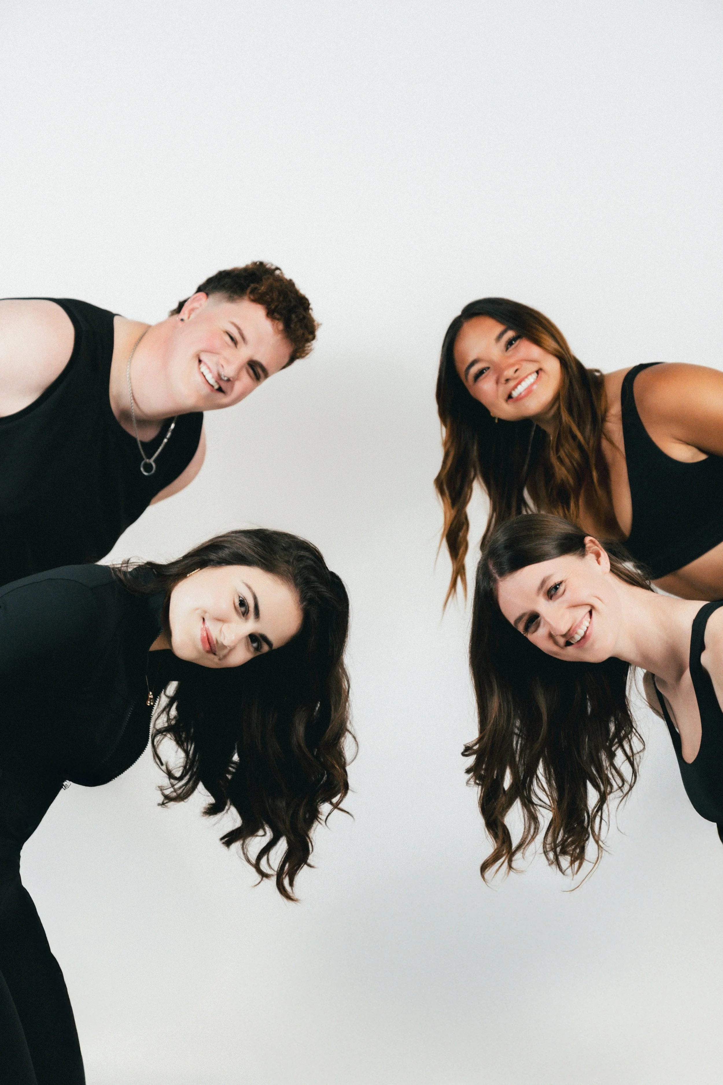Group of five young adults, three women and two men, smiling and leaning towards the camera in a circle against a plain white background, dressed in black tops.