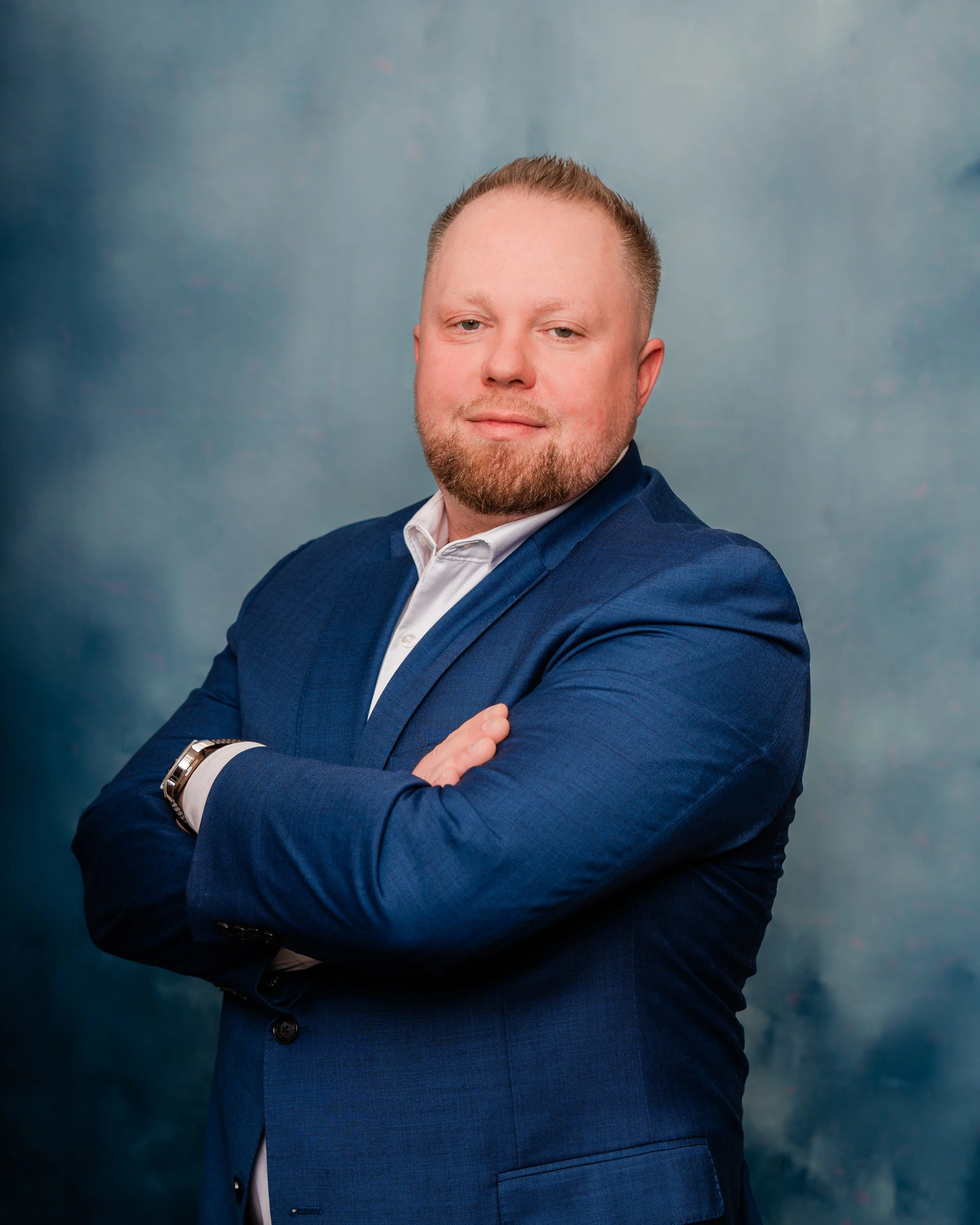 A man with a beard and short hair, wearing a blue suit and white shirt, standing with arms crossed in front of a cloudy blue background.