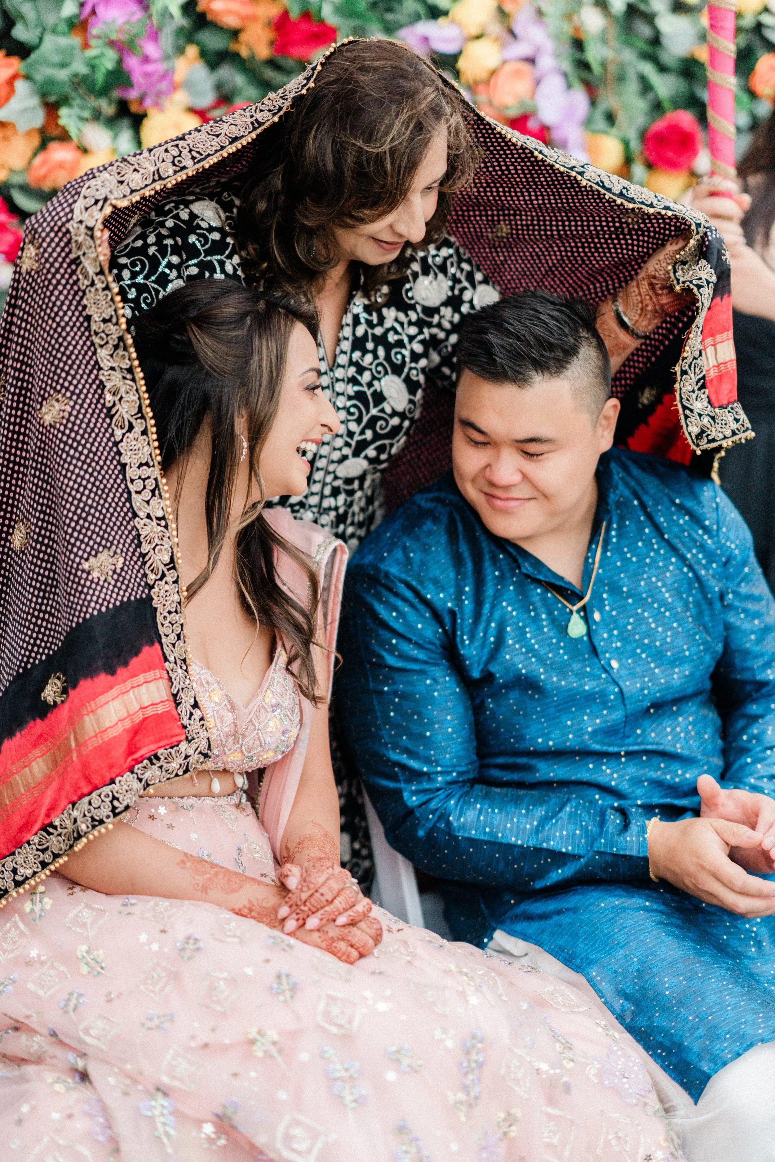 A woman dressed in traditional Indian attire is sitting with a man, both smiling, while an older woman, possibly a family member, stands over them, also smiling. The scene is decorated with colorful flowers in the background, indicating a festive cel