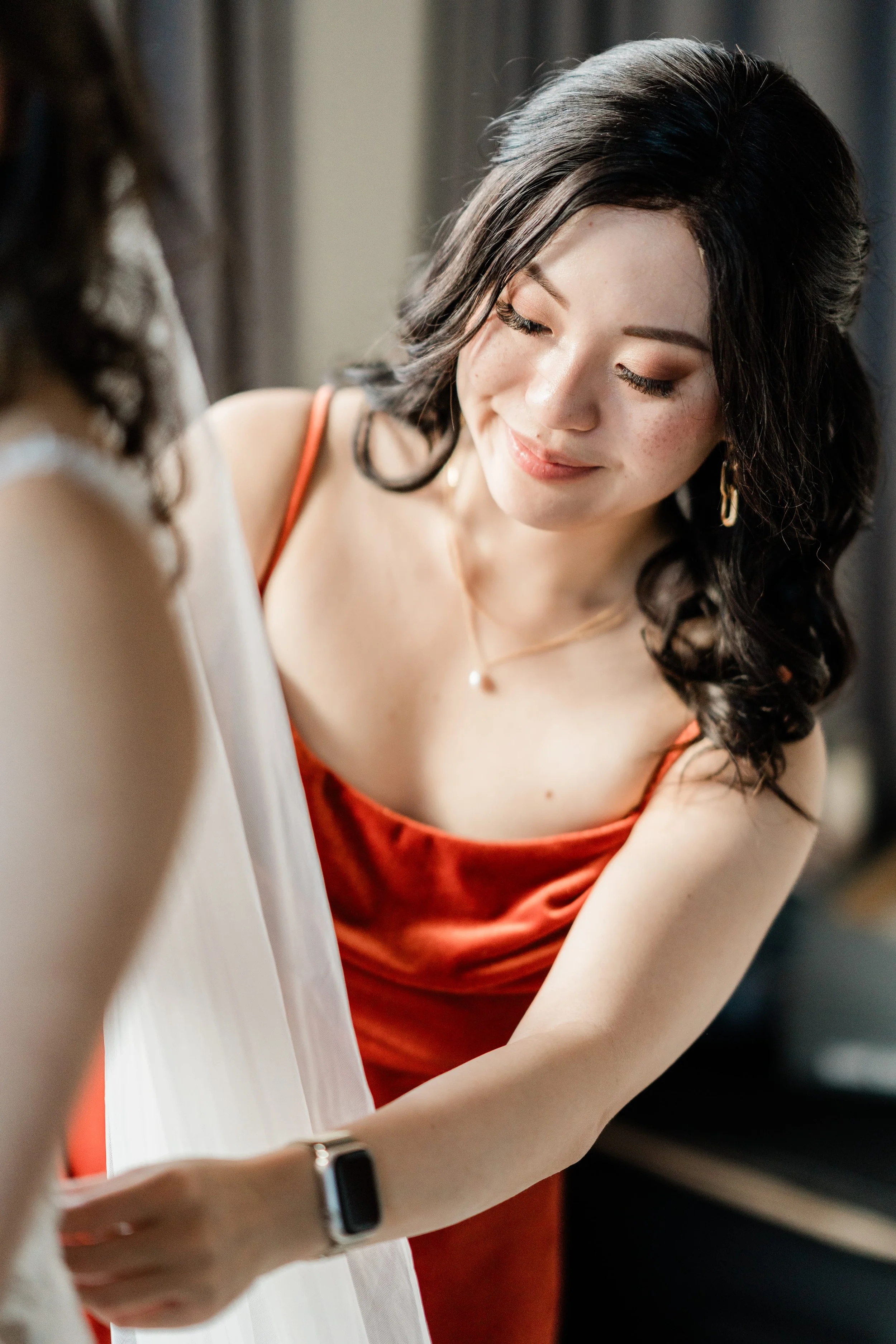 A woman in a red dress and gold jewelry, smiling softly and looking down, standing beside another person with blurred features, possibly adjusting something on her dress or outfit in a well-lit room.