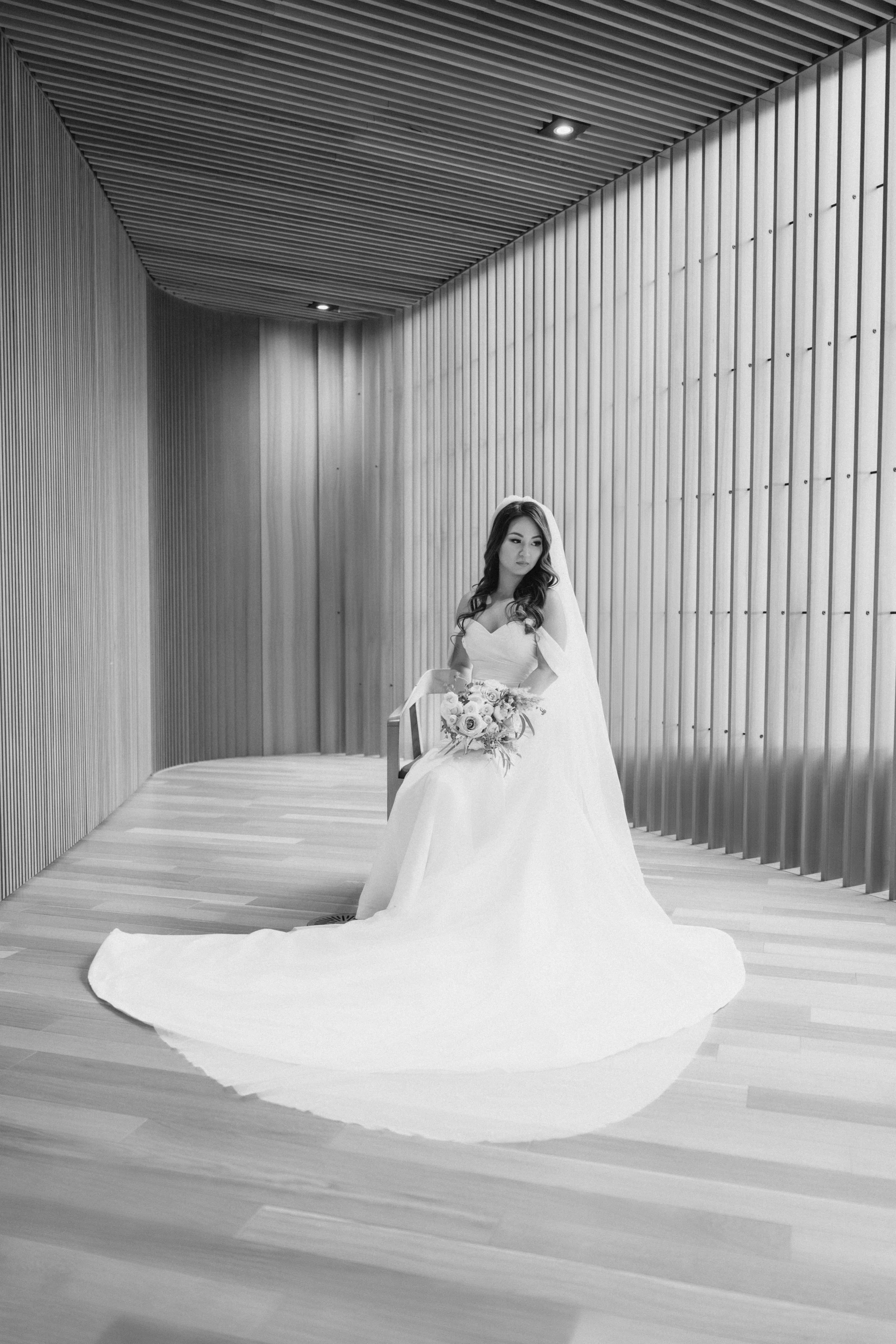 Black and white photo of a bride sitting on a wooden chair in a modern, wooden-paneled hallway, holding a bouquet, wearing a wedding dress and veil.