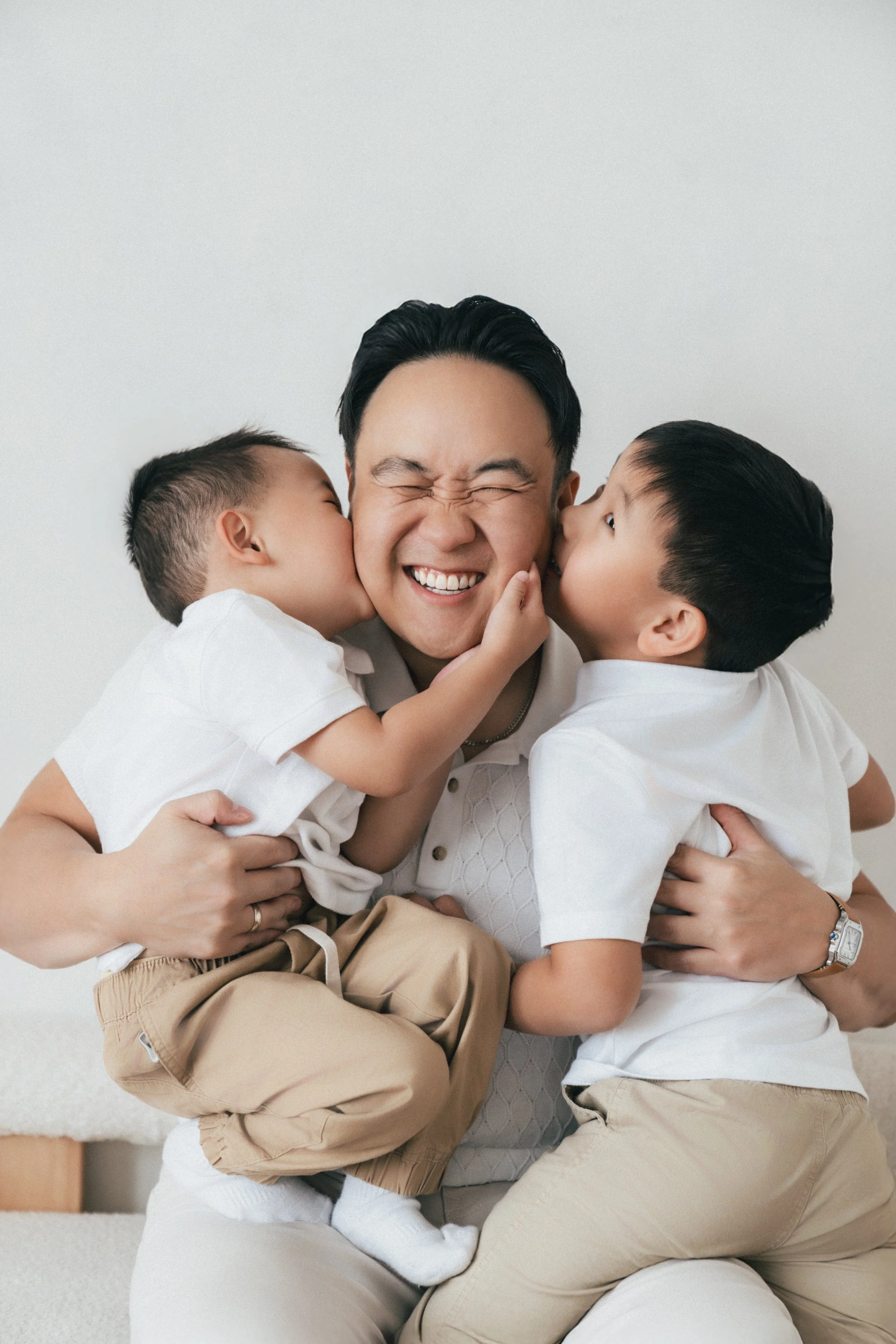 A father is being kissed on each cheek by two young children, both dressed in white shirts and beige pants, in a joyful, affectionate moment.