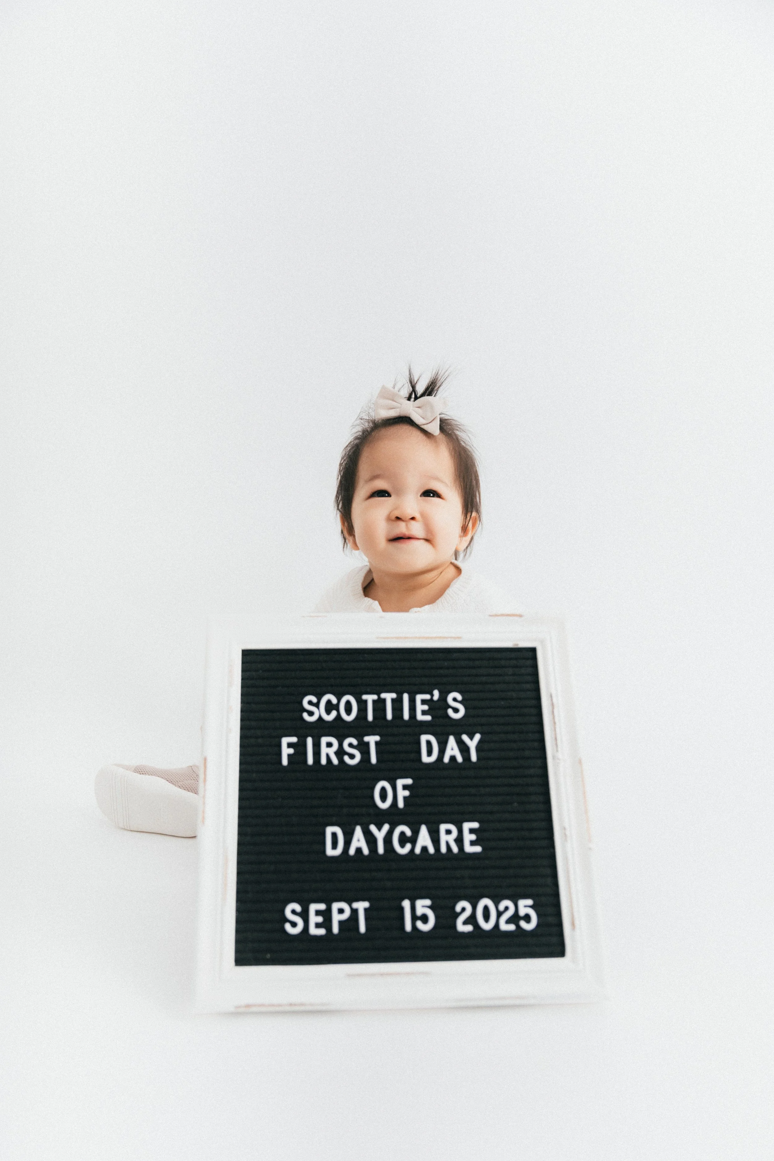 A smiling baby girl with a bow in her hair sitting behind a sign that reads "Scottie's first day of daycare September 15, 2025" against a white background.