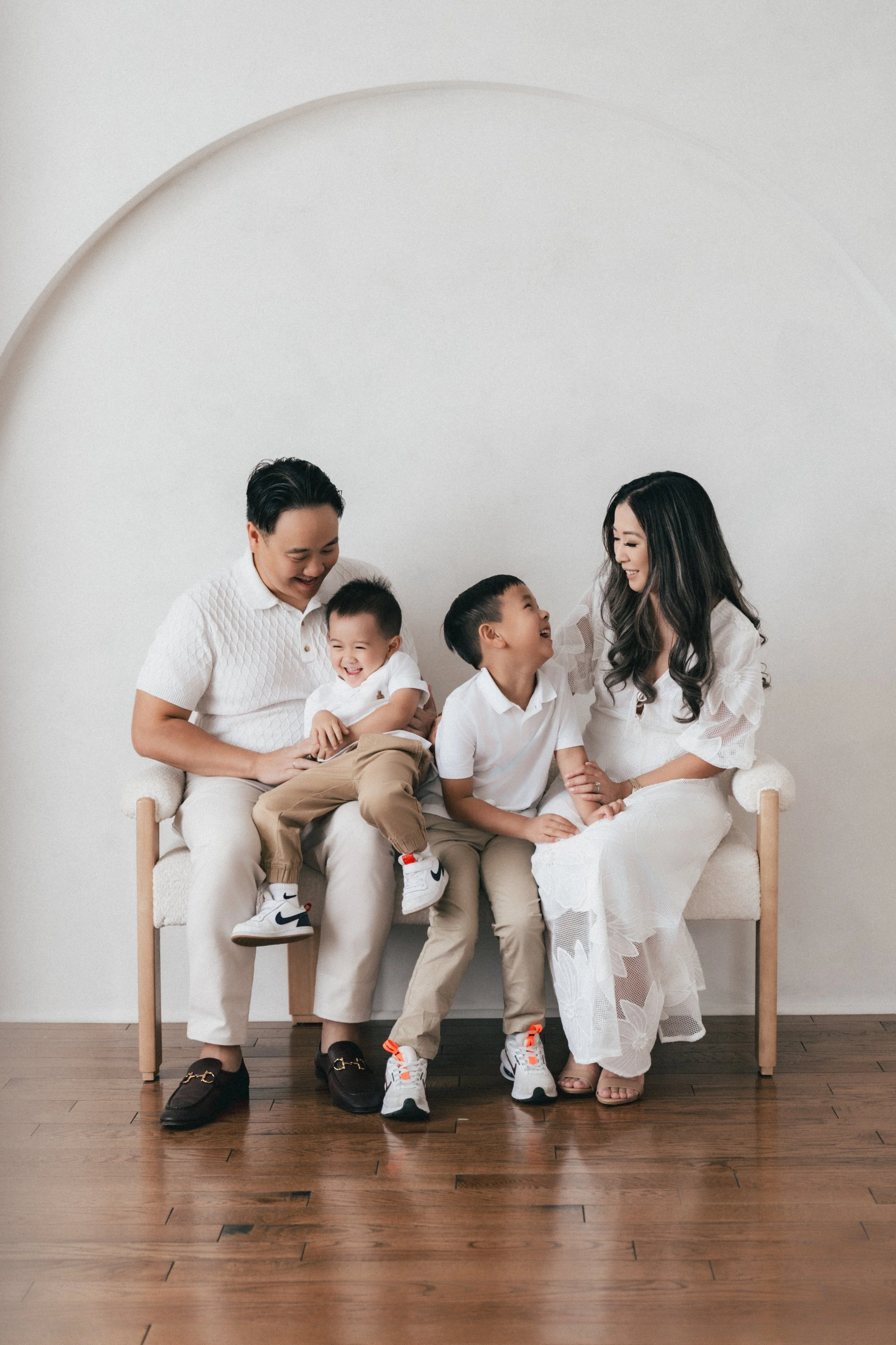 A family of four sitting on a white couch, laughing and smiling together in a minimalist room with wooden flooring.