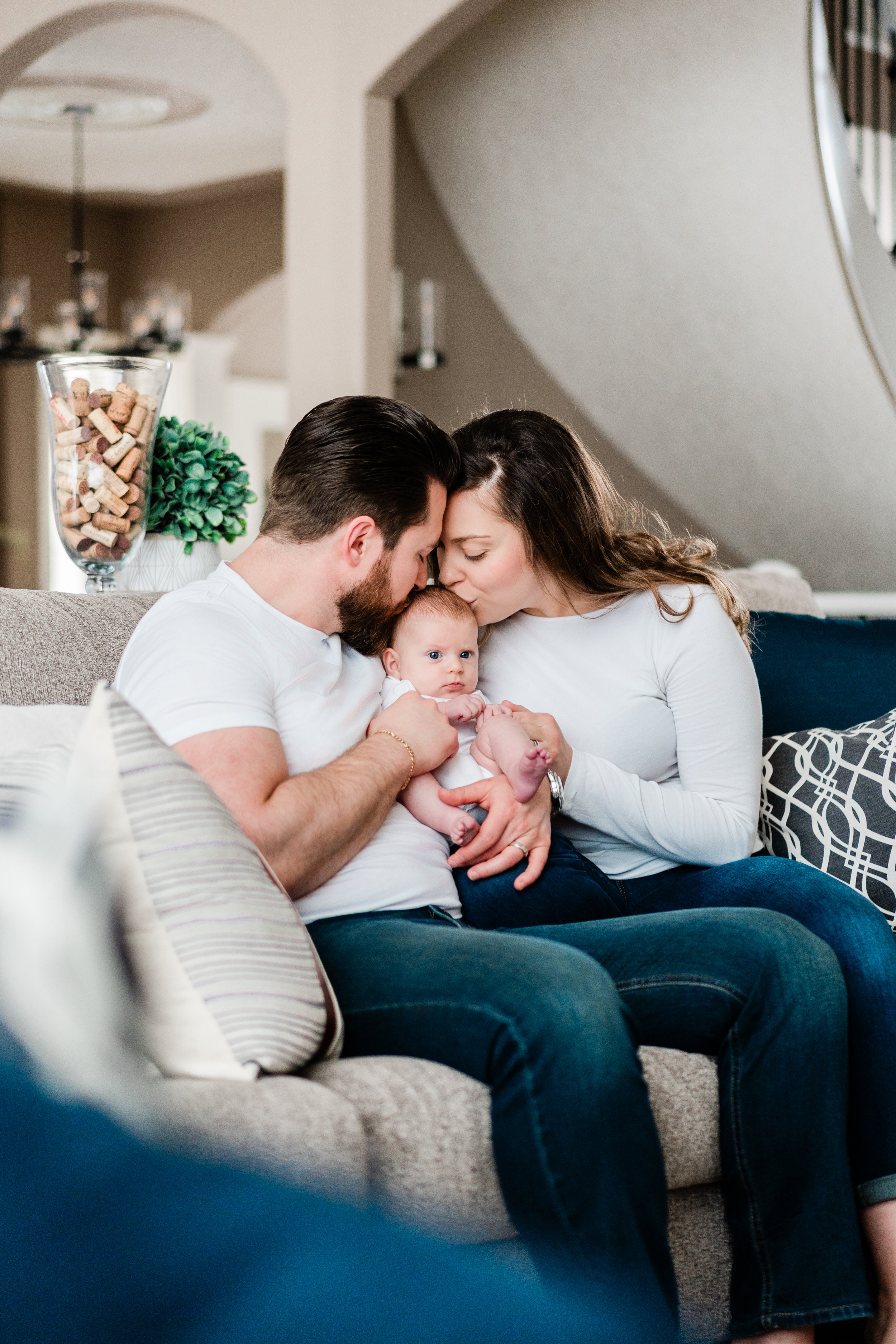 A young family sitting on a beige couch, the mother and father affectionately kissing their baby on the forehead.