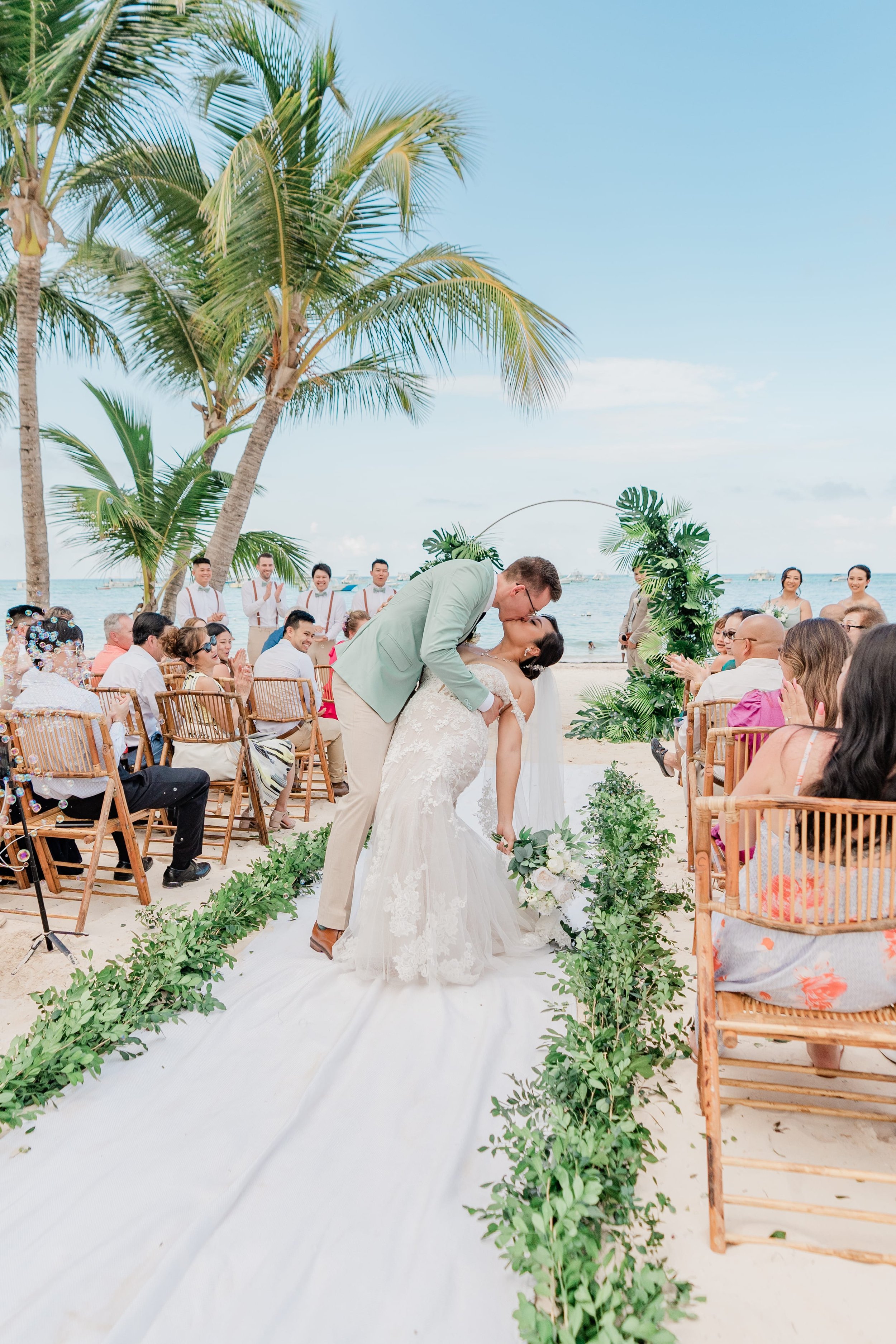 A bride and groom share a kiss at an outdoor beach wedding with guests seated on either side and tropical palm trees in the background.