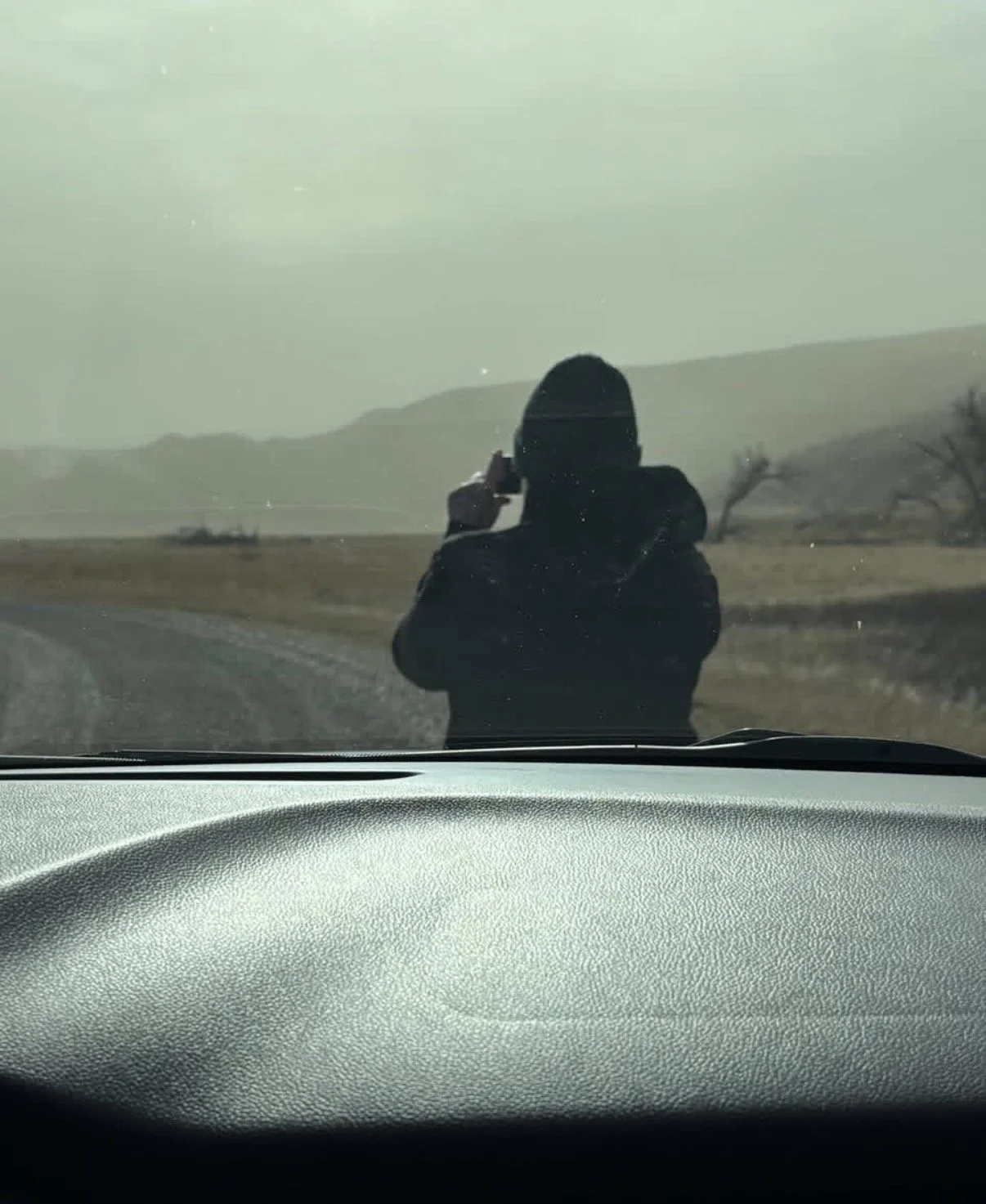 A person standing outside in a field, wearing a hooded jacket, taking a photo or video with a mobile phone, with hills and leafless trees in the background, viewed from inside a car.