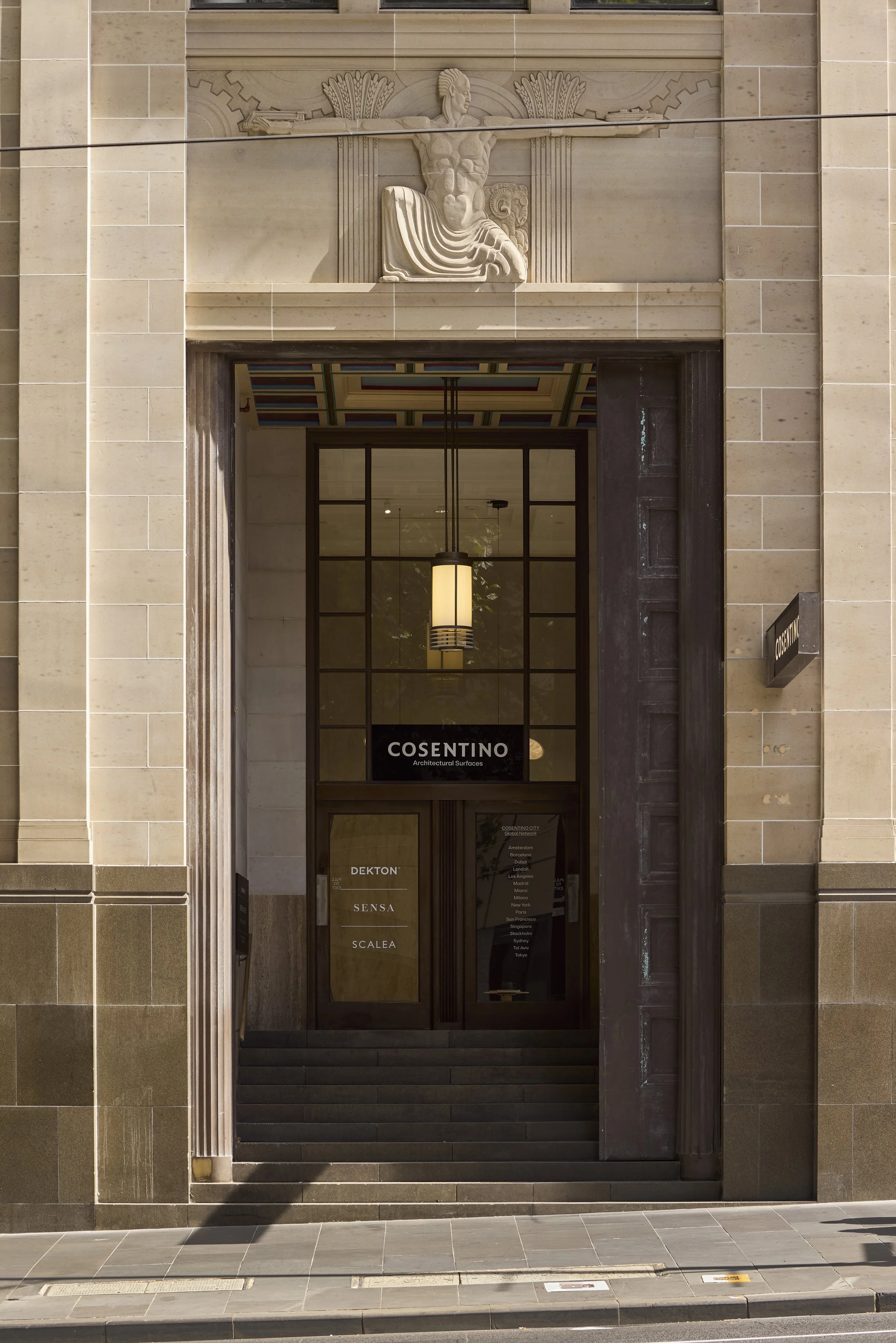 Entrance to a building with a carved stone relief of an ancient Greek figure with outstretched arms above the doorway, and a modern hanging light fixture inside. The building has beige stone exterior walls and a dark-colored door and frame.