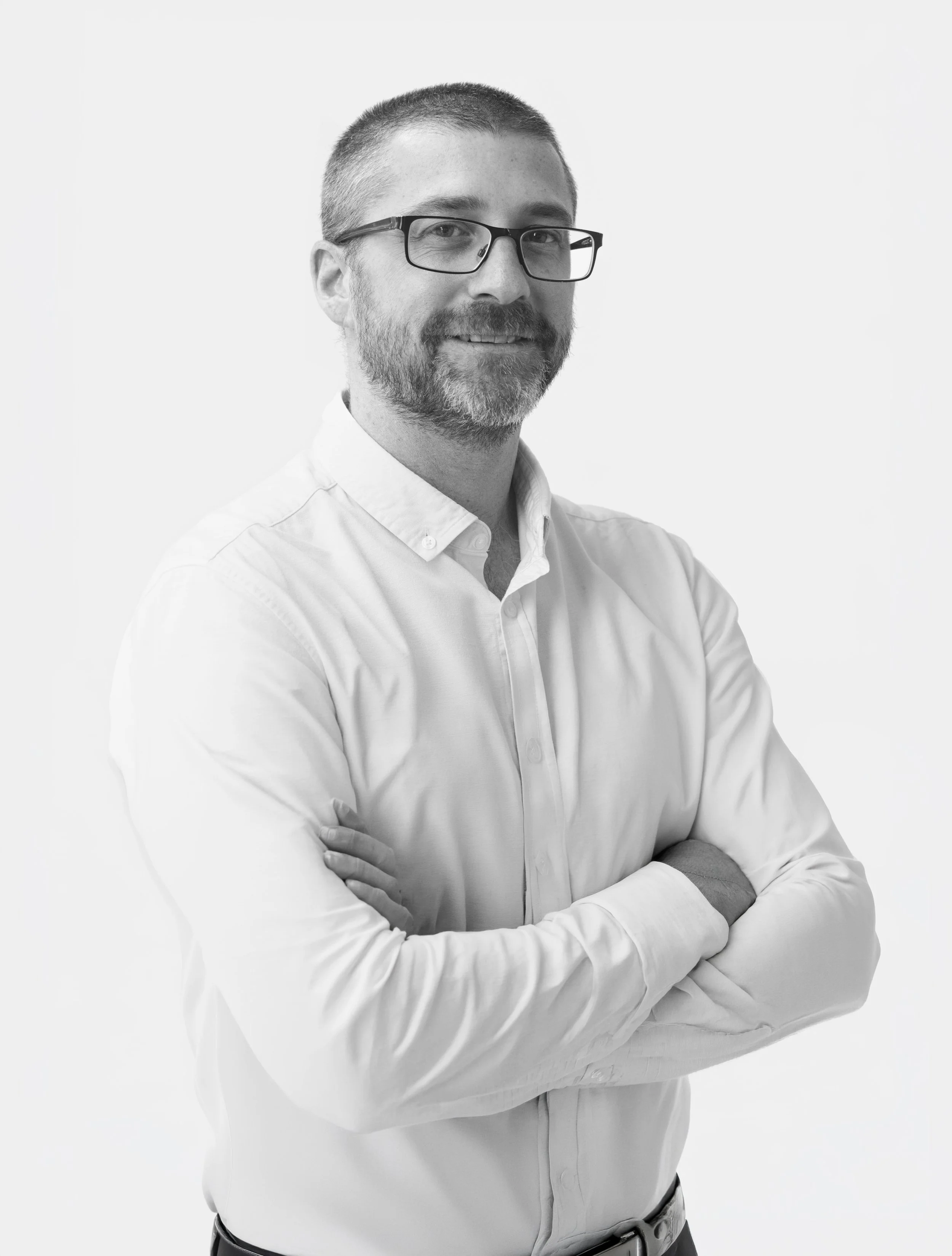 Black and white portrait of a man with glasses, a beard and short hair, wearing a light coloured shirt, standing with arms crossed against a plain white background.