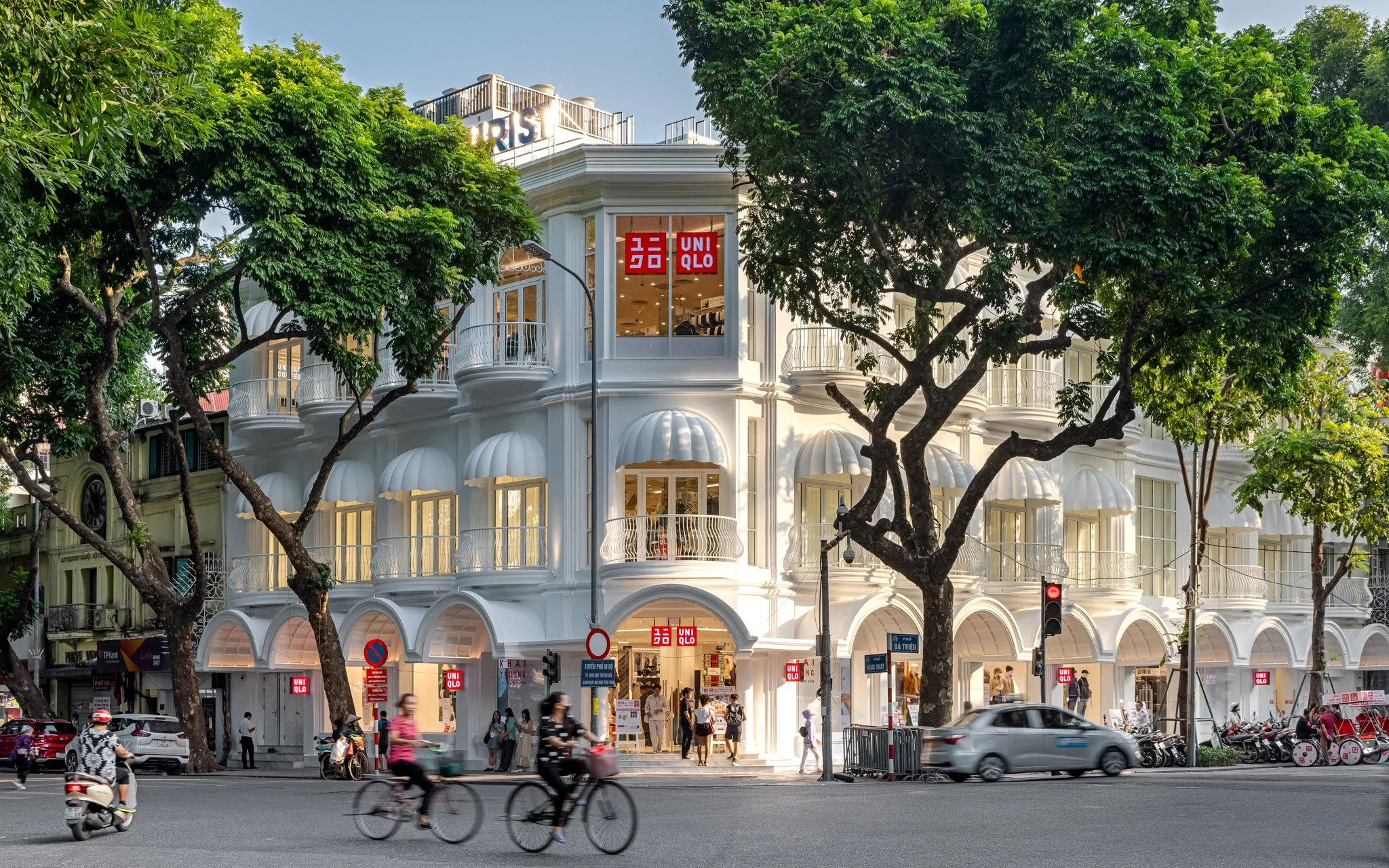 A busy city street with a white, multi-story building featuring balconies and large windows, partially obscured by green trees. The building houses a UNIQLO store, visible through red signs with white text. Pedestrians and cyclists are on the street, with cars and motorbikes passing by.