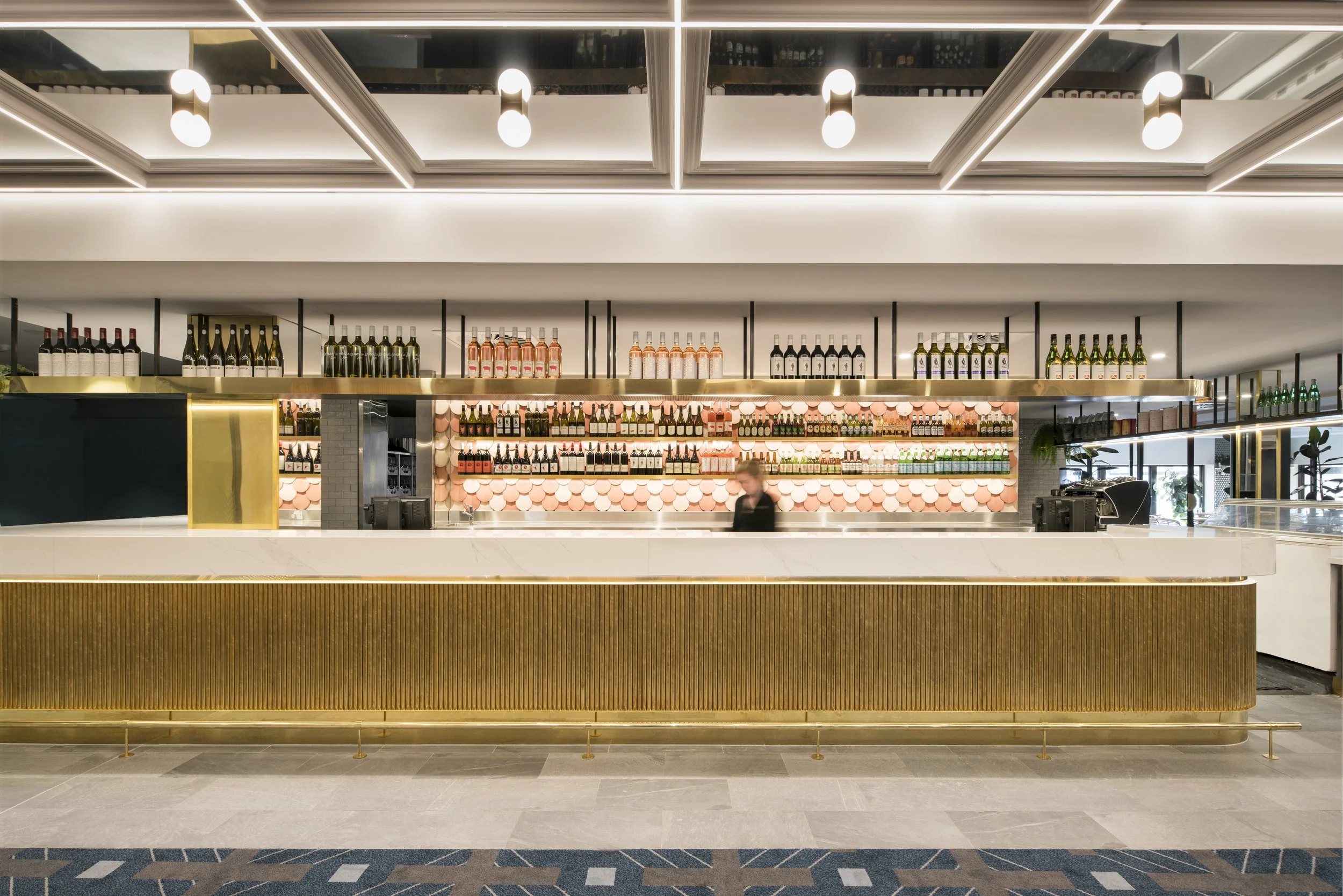 Modern bar counter with shelves of wine bottles, bottles of sparkling water, and decorative pink and black circular accents. The area is well-lit with ceiling lights and has a clean, stylish design.