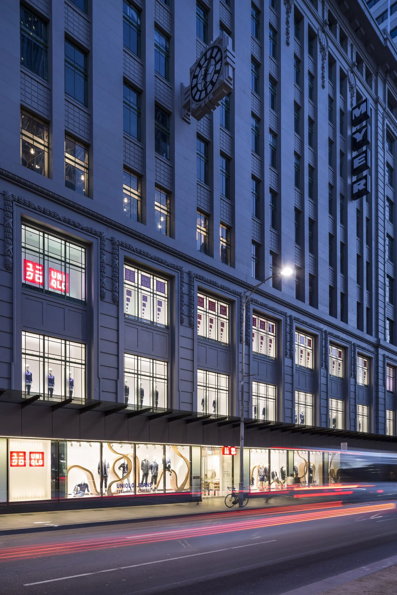 A multi-story building with a clock on its facade, featuring large windows with mannequins dressed in clothing inside. The storefront displays the UNIQLO brand logo and advertisements for clothing, with nighttime lighting illuminating the street scene, including streaks of light from passing vehicles and a bicycle parked outside.