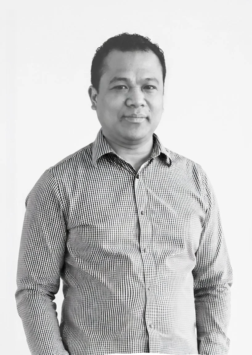 Black and white photograph of a man with short, dark hair wearing a checkered button-up shirt, standing against a plain white background.